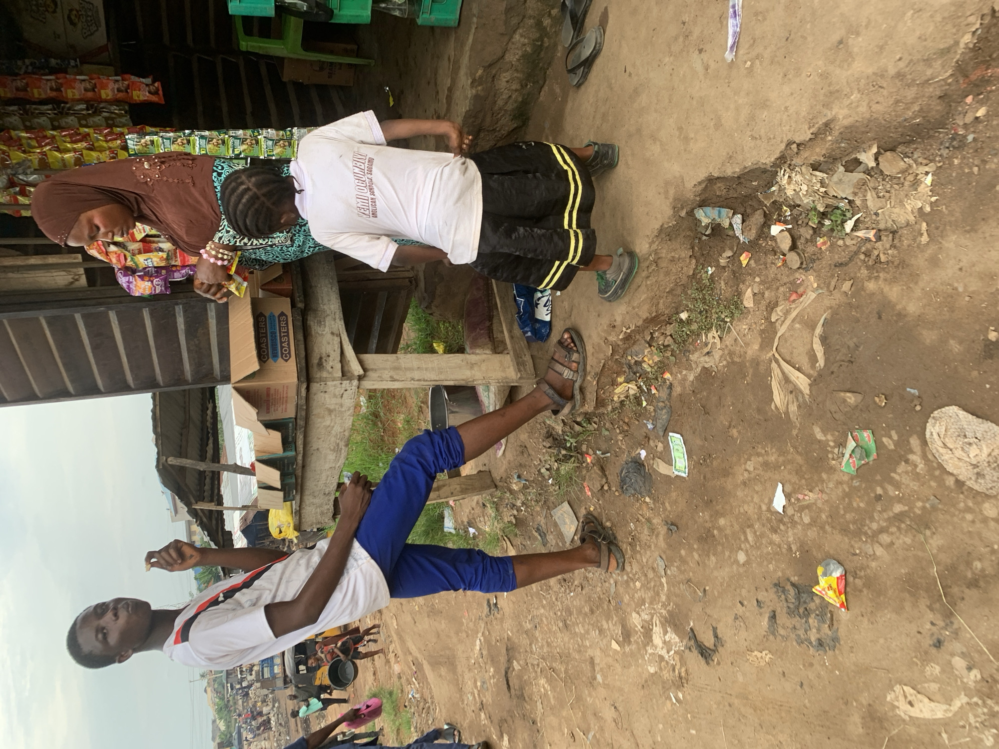 A young Black male wearing a white t-shirt and blue pants stands on a dirt path in front of a wooden stall in Shagamu, Nigeria. He is holding a piece of food in his right hand and looking forward. Behind him, a Black female wearing a brown hijab and a patterned dress is interacting with a young Black female wearing a white t-shirt and a black skirt. The stall has shelves stocked with goods, and boxes are stacked on a table. The ground is littered with trash and debris. In the background, there are other people, vehicles, and buildings, suggesting a busy market or street scene. The overall environment appears to be urban and somewhat unkempt.