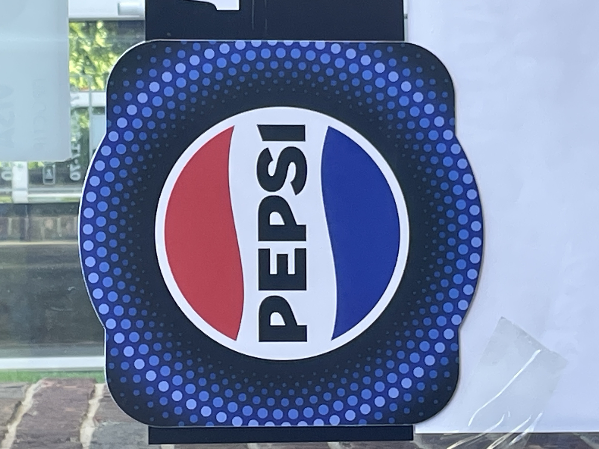A Pepsi logo sign is attached to a glass door or window. The sign is a square with rounded corners and a scalloped bottom edge. The Pepsi logo, a circle with red, white, and blue sections and the word "PEPSI" in white text, is centered on the sign. The background of the sign is a dark blue with a pattern of lighter blue dots that fade from light to dark. Behind the sign, through the glass, an outdoor scene with trees and foliage is visible, along with an interior scene featuring a brick wall and part of another sign. The sign appears to be new and in good condition.