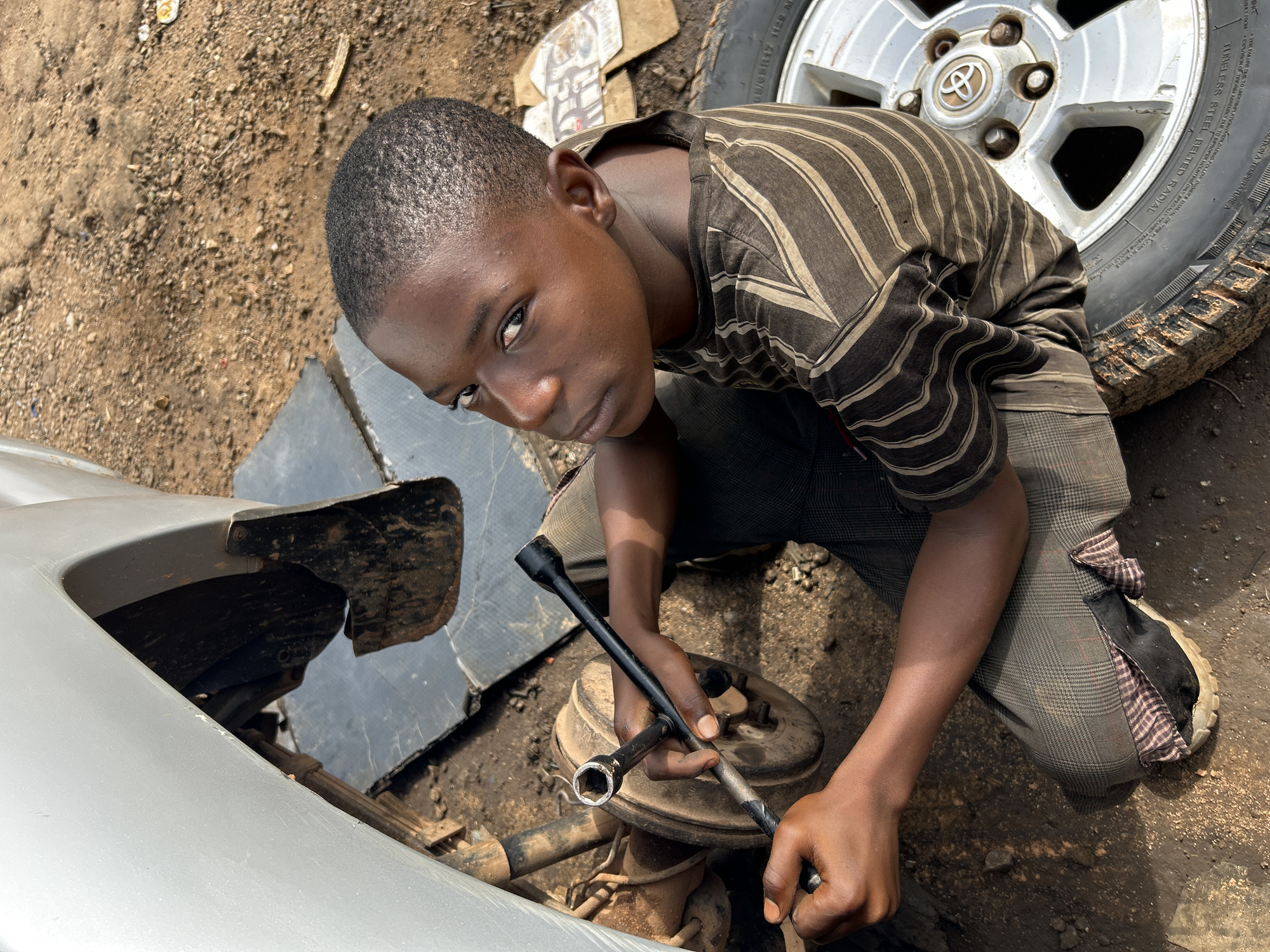 In Barikin Jirga Summa, Nigeria, a young boy kneels on the ground, engaged in repairing a car. He is focused on the car's wheel assembly, holding a wrench in his hands as he works on the brake drum. The surrounding environment is dusty and consists of dirt and debris. A silver car, partially visible, is the central subject of the repair. The boy is dressed in a striped t-shirt and patterned trousers, with a worn shoe on his left foot.