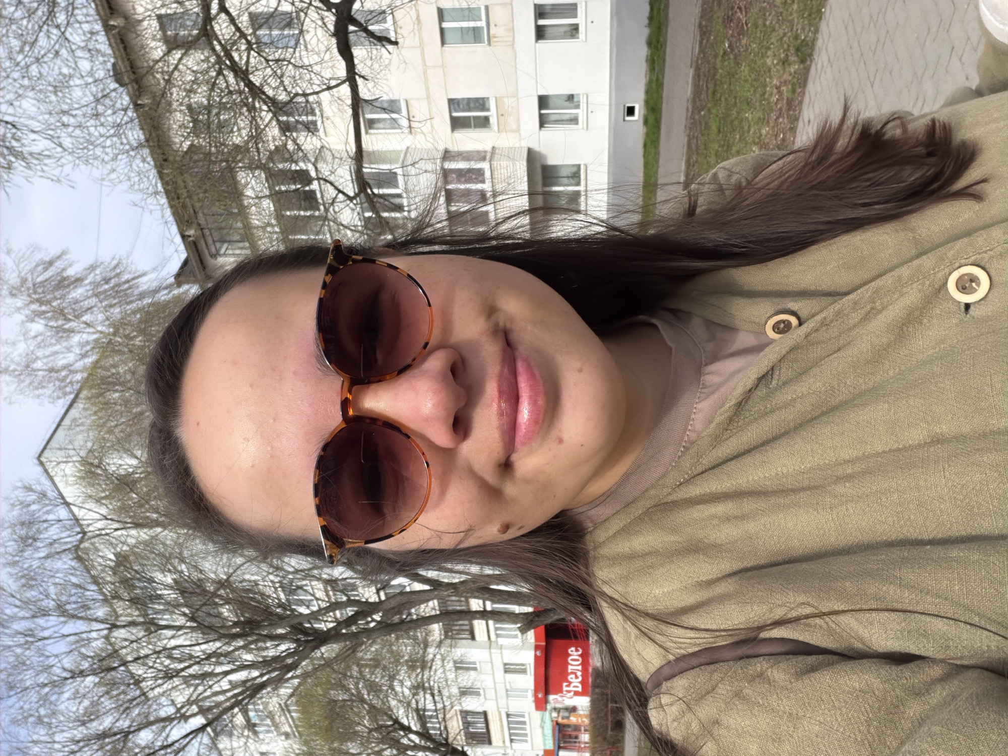 A woman wearing sunglasses and a green shirt is outdoors in Chaikovsky, Russia. Behind her is a building and trees. A sign with the word "Белое" (Beloe, meaning "White" in Russian) is visible in the background. The photo is taken from a slightly low angle. The focus is primarily on the woman and her immediate surroundings.