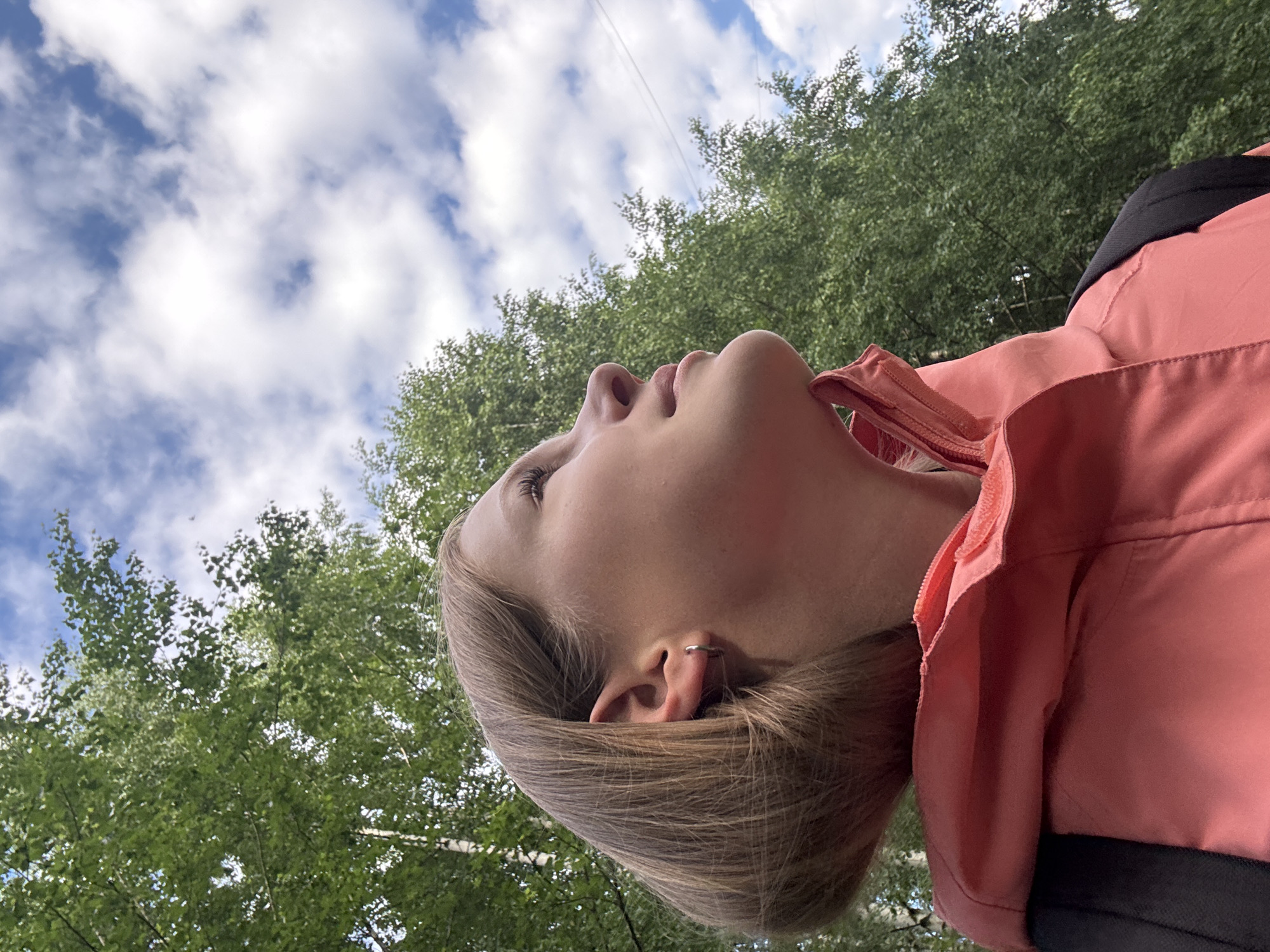 This is a vertical, low-angle shot of a young person, seen from the neck up, as they look upwards towards the sky. The subject has light brown hair and is wearing a pink jacket with a black collar peeking out from underneath. They are wearing a small silver hoop earring in their visible ear. The background consists of a bright blue sky with scattered white clouds, and the lush green foliage of trees. The image appears to be taken outdoors on a sunny day, with the upward angle suggesting the subject is lying down or looking up from a low vantage point. The overall mood is relaxed and contemplative, with the subject's gaze directed towards the open sky and greenery. The specific location, Chaikovsky, Russia, is mentioned as context but is not visually identifiable from the image itself. There is no visible text in the image.