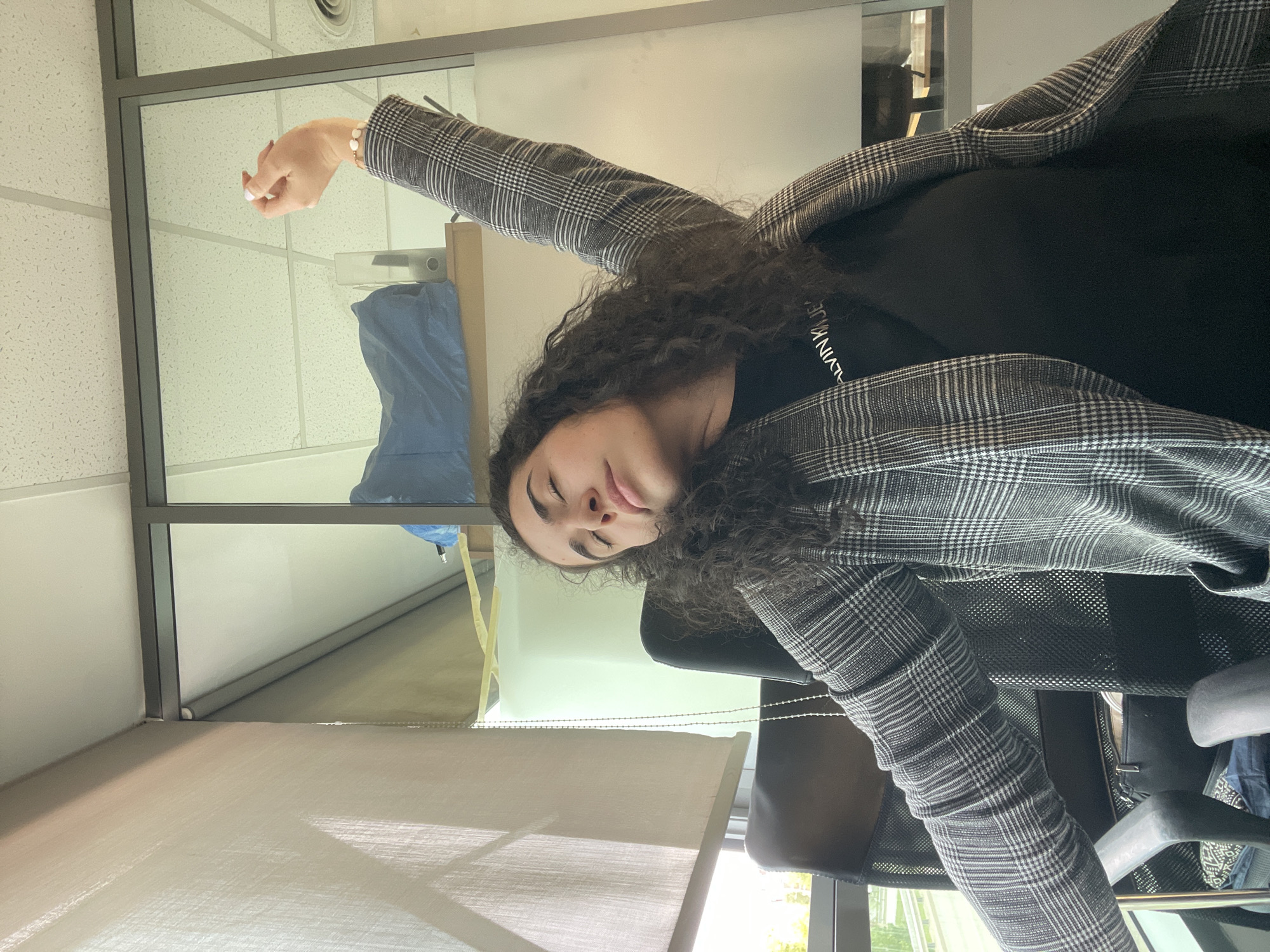 The image shows a woman with curly dark hair, wearing a black top with "Calvin Klein" visible on the neckline and a grey and white plaid blazer, sitting in an office chair. She is in what appears to be an office setting, likely inside an office building. Her right arm is raised, and she has a bracelet on her wrist. Light can be seen through the window, suggesting it is daytime.
It's impossible to determine whether the photo was taken in Sofia, Bulgaria, or even definitively that it was taken indoors.