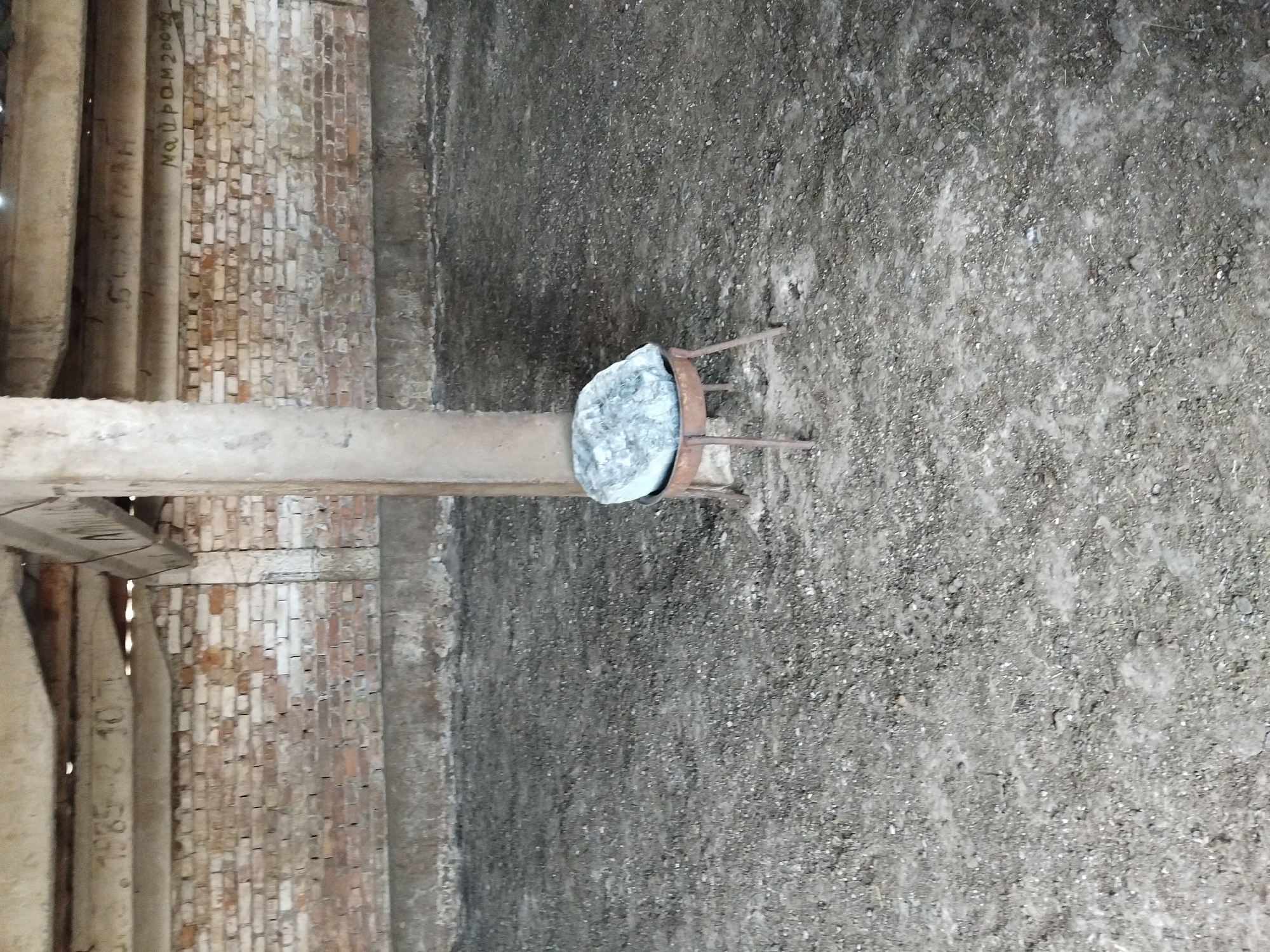 A rough, textured stone sits on a small, metal stool in the center of a dusty, gray-brown dirt floor. Behind the stool, a brick wall, constructed with varying shades of red and white bricks, stretches across the frame. Above the wall, a concrete beam is visible, bearing faint markings. A thick, concrete pillar stands to the left of the stool, extending upwards towards a wooden ceiling structure. The overall scene appears to be an interior space with a rustic, possibly abandoned, aesthetic.