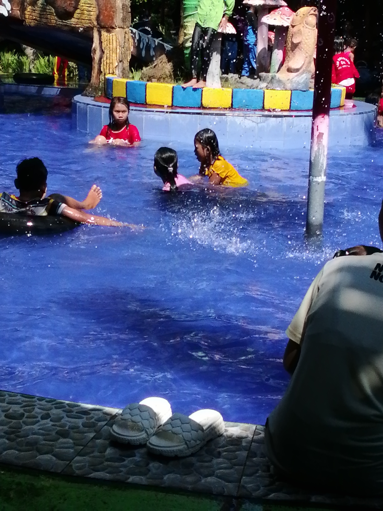 The image shows a pool area, likely at a water park, with several children and adults present. The pool is filled with blue water, and there are some water features, including a decorative structure with mushroom-like elements and a fish sculpture. Some of the children are in the water, playing and splashing. In the foreground, a person's back is visible, wearing a light-colored t-shirt. Two pairs of sandals are placed on the stone edge of the pool. In the background, there are more people, some appearing to be adults supervising. The environment suggests a sunny day with water activities taking place. The location is identified as Tegalwangi, Indonesia.