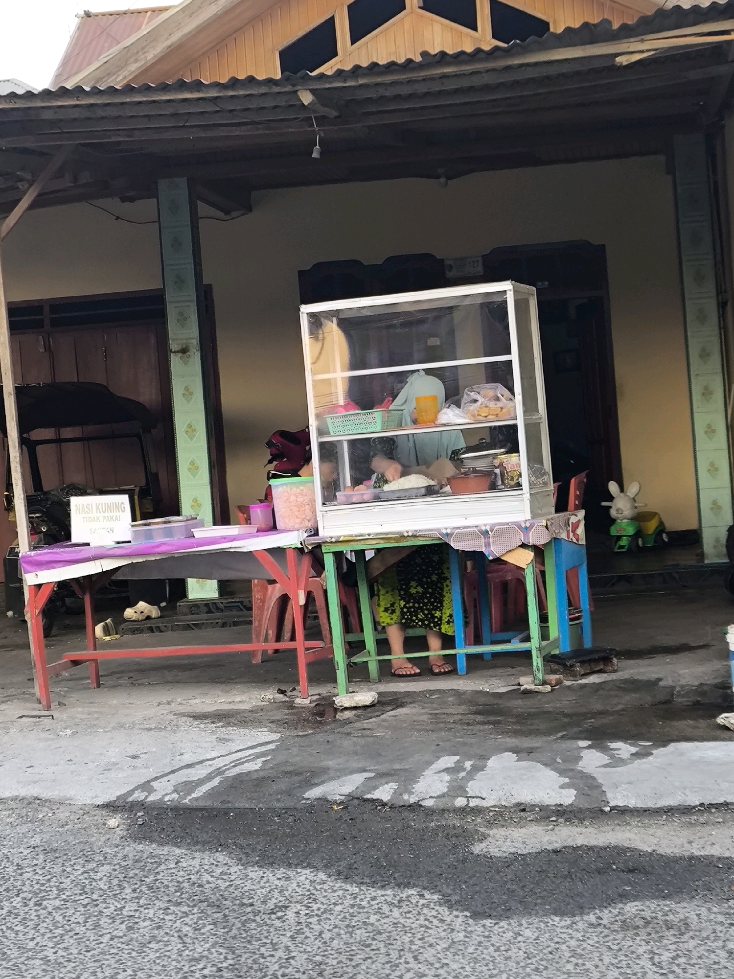 The image shows a street scene in Gorontalo, Indonesia, likely a food stall. A woman is seen behind a glass display case, which contains food items. In front of the display case, a table is set up with various containers and a sign that reads "NASI KUNING TIDAK PAKAI SAMBAL". A toddler's toy rabbit ride-on is visible in the background. The stall appears to be set up in front of a building with a corrugated metal roof. The ground is paved with asphalt and concrete. The overall impression is that of a typical roadside food vendor.
