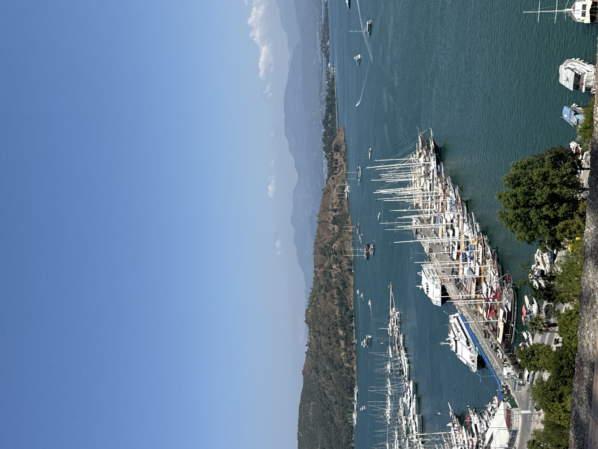This image captures a panoramic high-angle view of the bustling harbor and bay of Fethiye, Turkey, under a clear, bright blue sky with minimal scattered cirrus clouds on the distant horizon. The foreground features a rough-hewn stone parapet with dense green foliage, including trees and bushes, indicating the photograph was taken from an elevated vantage point.
The expansive turquoise-to-deep-blue waters of the bay dominate the mid-ground and background. Numerous vessels are present. In the lower-left and central portions, a large marina contains dozens of docked sailboats and yachts, identifiable by their prominent white masts and rigging, moored to multiple long piers. Several smaller motorboats are also interspersed among them.
Further into the bay, two large, modern white passenger ferries, one featuring a distinct blue stripe along its hull, are prominently docked at a substantial pier. Adjacent to these ferries, a cluster of traditional wooden gulets or tour boats, characterized by their multiple dark masts and classic design, are also moored. A road or paved area with several parked cars and low-rise white buildings runs parallel to this main pier.
Scattered across the open water of the bay are various other boats, including several motor yachts and smaller speedboats, some leaving visible white wakes, indicating movement. In the middle distance on the left, a large, tree-covered peninsula or hillside extends into the bay. In the far background, hazy blue-grey mountains define the distant skyline. Towards the bottom right of the image, two older, smaller boats are partially visible, one with a blue tarp covering it, and another white sailboat is docked further right. The scene is brightly illuminated, suggesting mid-day conditions.