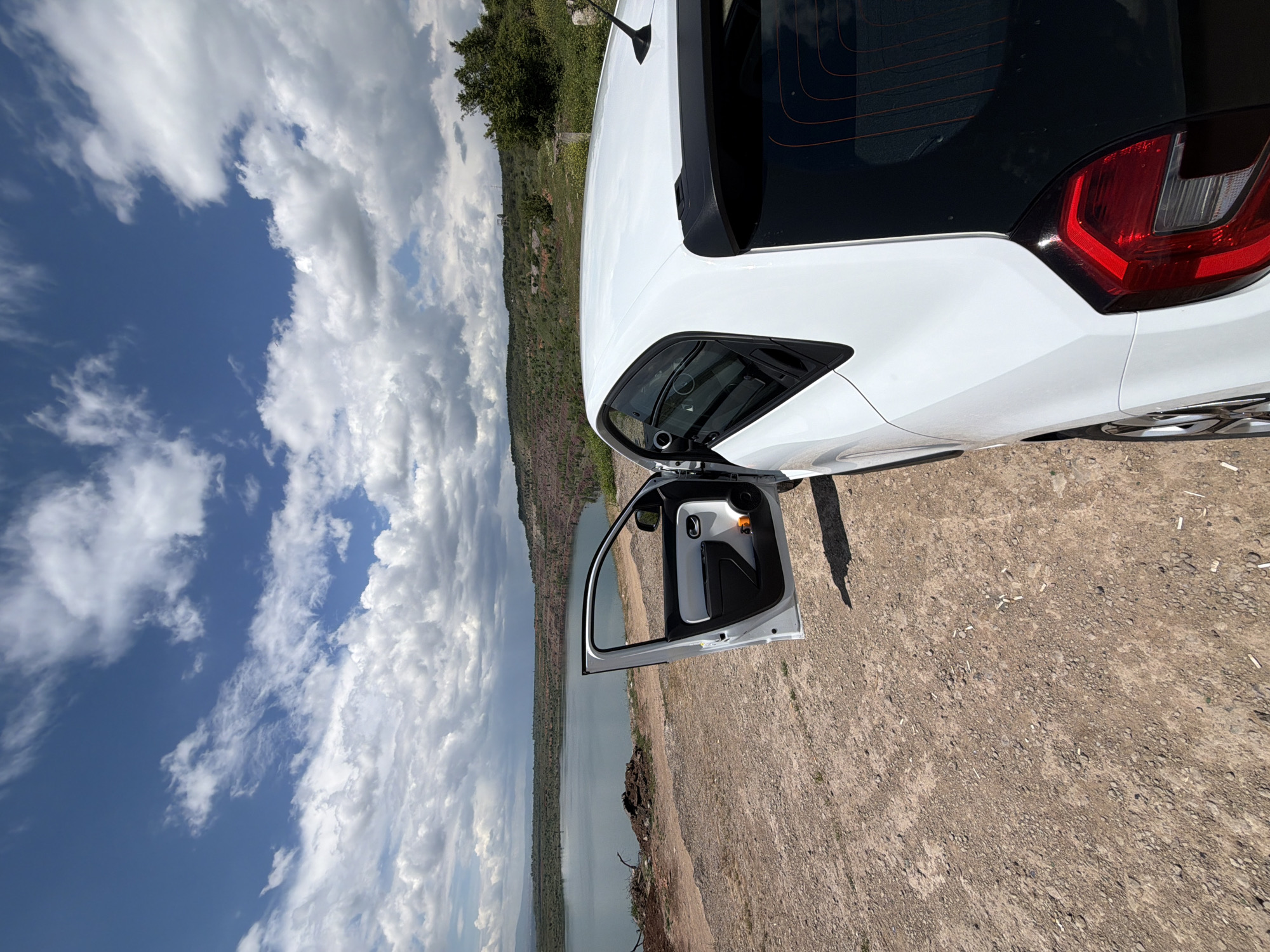 A white car is parked on a dirt area near a body of water under a cloudy sky. The car's driver-side door is open. The background includes hills and vegetation. The image appears to be taken at an elevated location. There is no information in the image to identify the exact location in Sofia, Bulgaria.