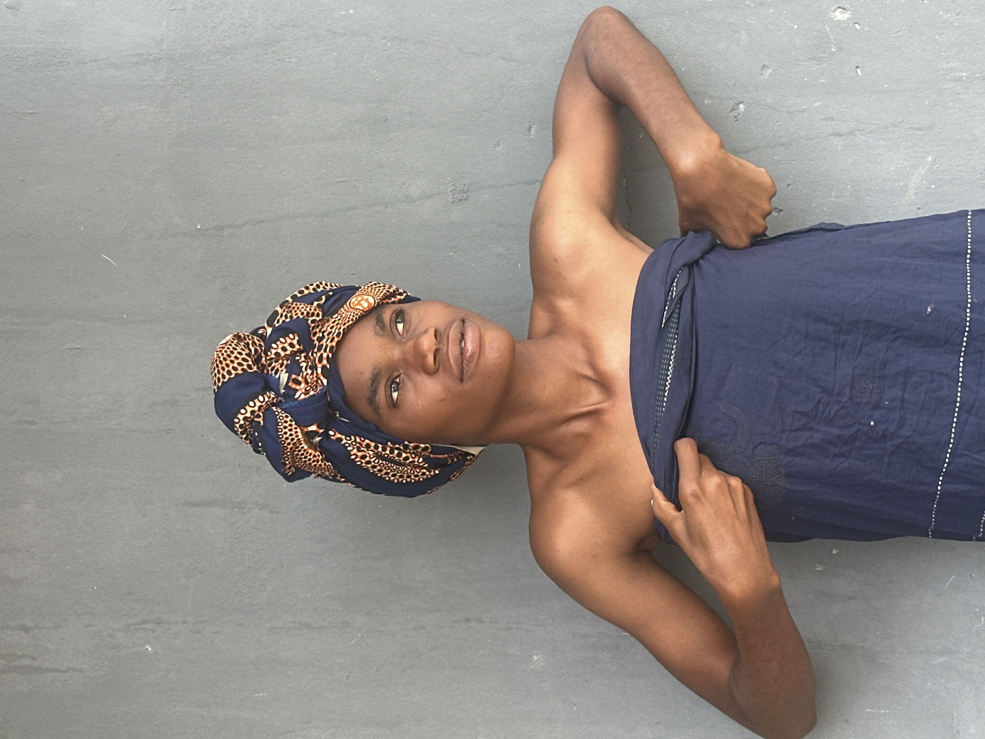 A young person with dark skin, wearing a blue and tan patterned headwrap and a navy blue cloth wrapped around their chest, stands against a grey concrete wall. The headwrap is tied in a bun on top of their head, with the pattern featuring blue, tan, and black circles and leopard-like spots. Their bare shoulders are visible. The person is looking to their right, with their lips slightly parted. Their arms are positioned as if adjusting the navy cloth, with their right hand holding the edge of the cloth at their chest and their left hand pulling it from the side. The wall behind them is rough and textured, with some white marks and what appear to be small cracks. The lighting suggests an outdoor setting, possibly during the daytime. The scene is static and the person is the sole subject. There are no other objects or people visible. The image appears to be a portrait taken in an outdoor urban environment, possibly in the city of Badagry, Nigeria, given the architectural style of the wall and the style of dress.
