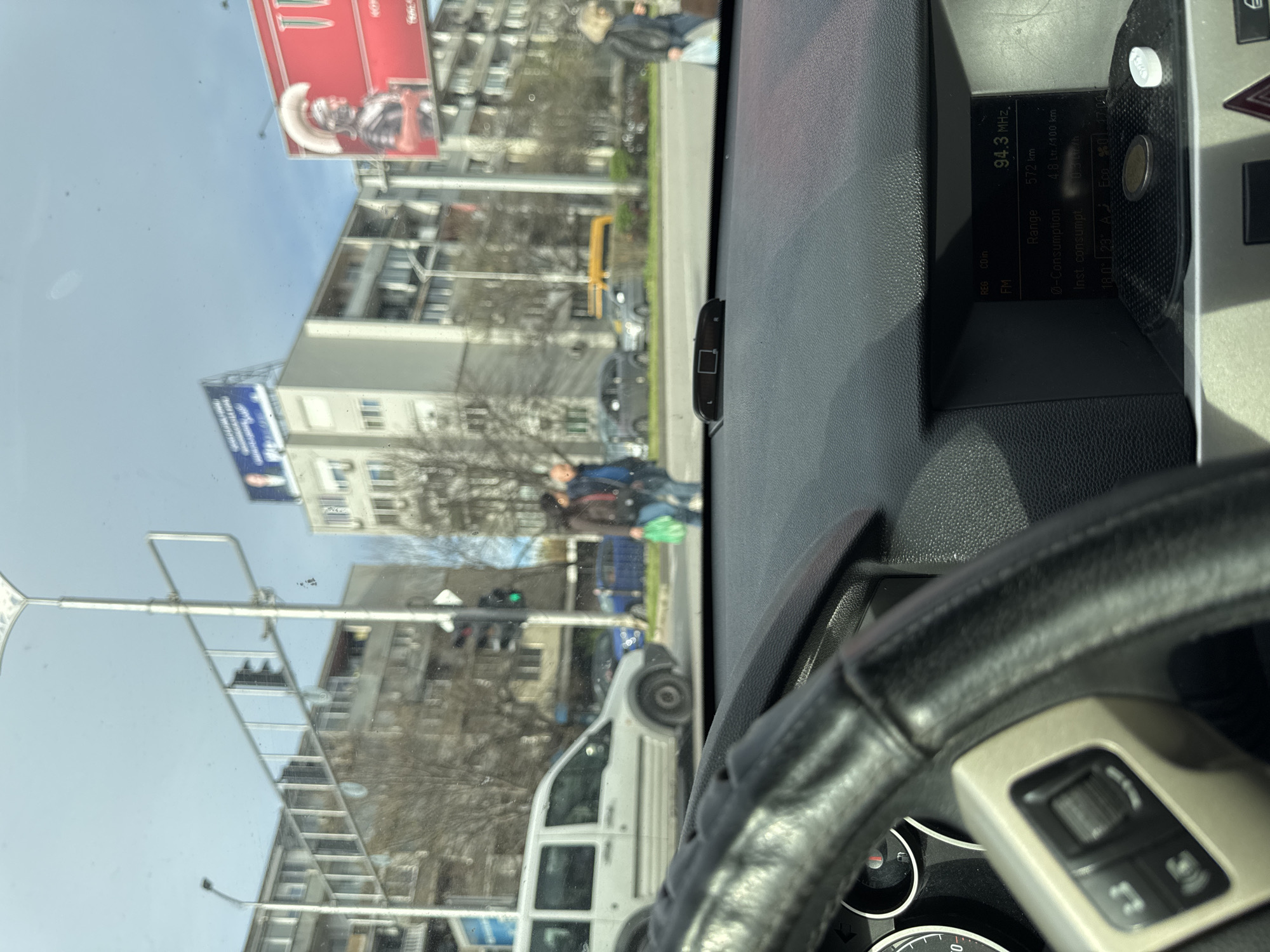 The image, taken from inside a car, shows an urban scene, probably in Plovdiv, Bulgaria. In the background, seen through the windshield, are apartment buildings, street lights, and billboards. The billboards feature images of a gladiator-like figure in red and what appears to be a political advertisement. People are visible on the street, including a couple walking. A white van is also visible on the street. Inside the car, the steering wheel is in focus in the foreground. The car's dashboard is visible, displaying information about the car's fuel consumption and range. It shows a radio frequency of 94.3 MHz. A small coin rests in a compartment in the dashboard. The weather appears to be clear and sunny.