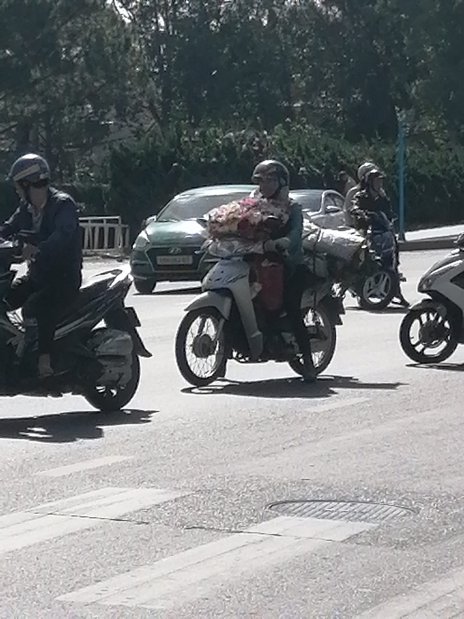 In Da Lat, Vietnam, a woman on a scooter, wearing a helmet, carries a bouquet of flowers. She is in the middle of the road, with a green car in the background and another scooter to her right. Trees line the street, and sunlight filters through the foliage. The scene appears to be a typical day in the city.