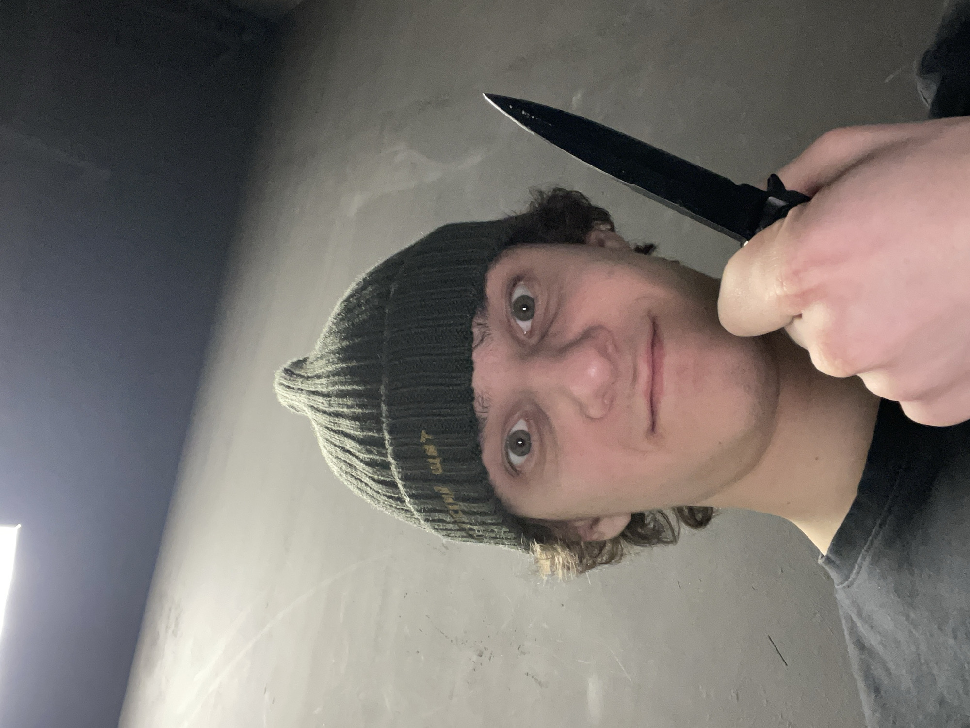 A person wearing a dark-colored beanie with gold-colored text is holding a black knife up to their face. The person has fair skin, light brown hair, and green eyes. The background appears to be an indoor setting with gray walls. The location cannot be determined to be Krasnoyarsk, Russia, or any other location, based on the visual information available in the image.