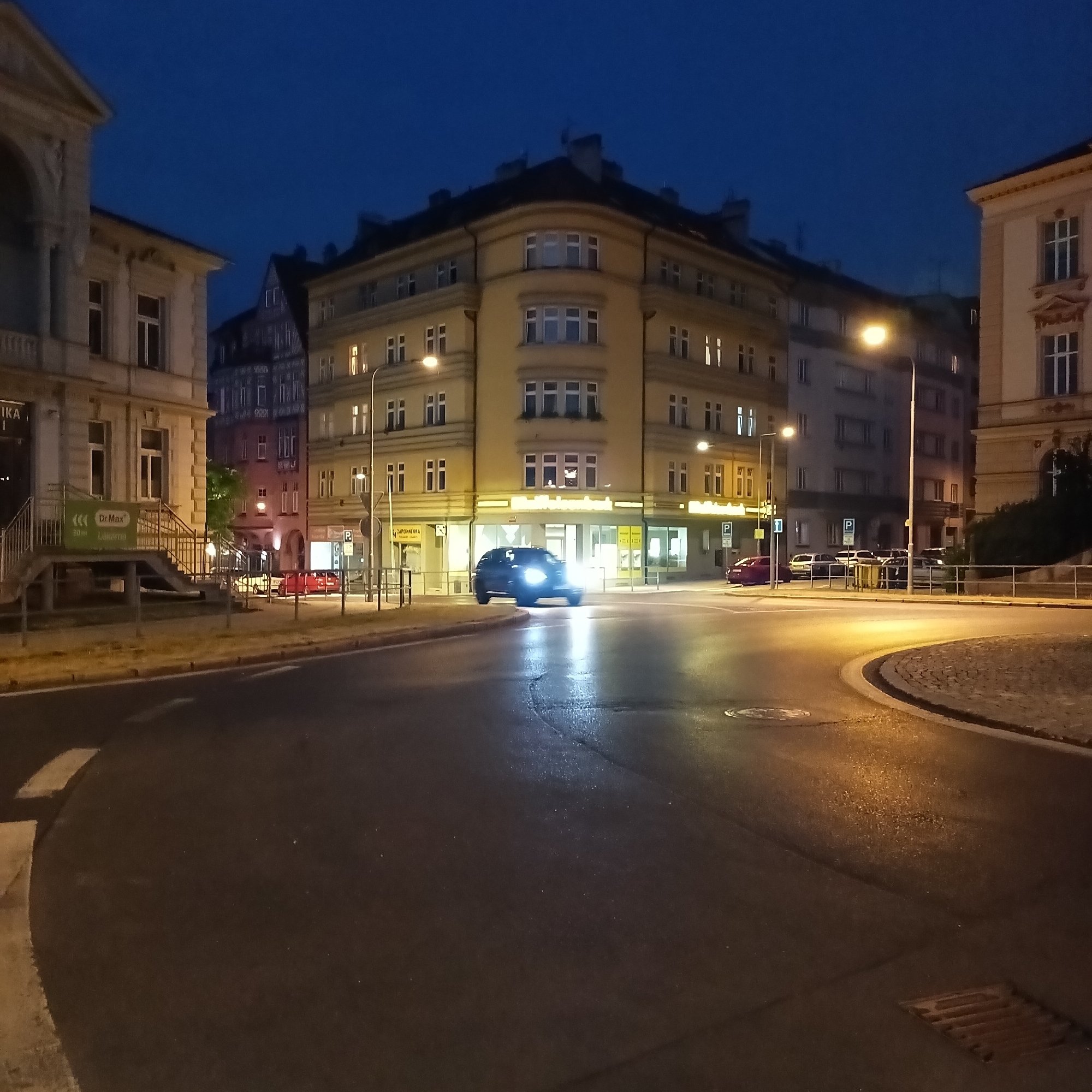 The image depicts a nighttime street scene in Cheb, Czechia, specifically at the intersection of Nádražní Street and Dlouhá Street. A dark-colored car is in motion, its headlights illuminated, turning at the intersection. Surrounding the intersection are several multi-story buildings with illuminated windows, suggesting residential or commercial occupancy. A street lamp illuminates the scene from the left side of the frame. The road surface is asphalt, appearing wet and reflective from the lighting. A pedestrian crossing is visible on the road. To the left, a building with classical architectural elements is partially visible, featuring columns and an arched entryway. Further left, a metal staircase leads up to a building entrance with a green sign that reads "Dr.Max Lekarna". On the right side of the intersection, parking signs are visible. The sky above is a deep, dark blue, indicating nighttime.