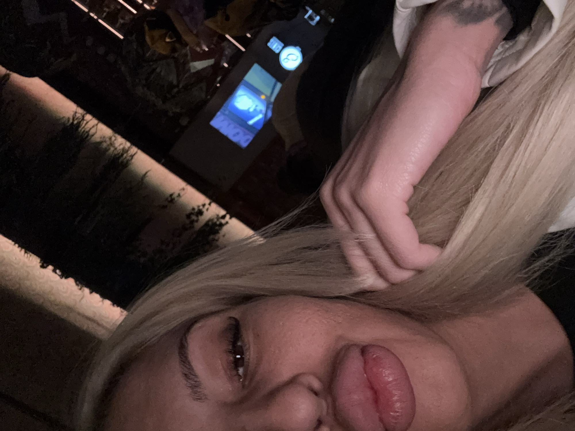A close-up, angled shot shows a woman with blonde hair, partially obscuring the background. Her face is prominently displayed, with her tongue extended. In the background, there's a blurred setting of what appears to be a dimly lit interior, potentially a restaurant or bar. A digital display, possibly from a cash register or a point-of-sale system, is partially visible. The location is not discernible as Plovdiv, Bulgaria, based solely on the visual information.