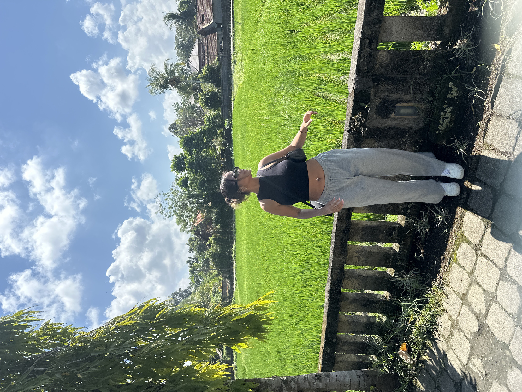 A young woman stands in front of a vibrant green rice paddy field under a bright blue sky with fluffy white clouds. She is wearing a black crop top, grey sweatpants, and white sneakers. Her hair is pulled up in a bun, and she is wearing sunglasses. She has a small black purse slung over her shoulder. The rice paddy is bordered by a low stone wall and lush vegetation. In the background, traditional Balinese houses with red tile roofs are visible amongst trees. The scene appears to be in Ubud District, Indonesia, known for its picturesque rice terraces.