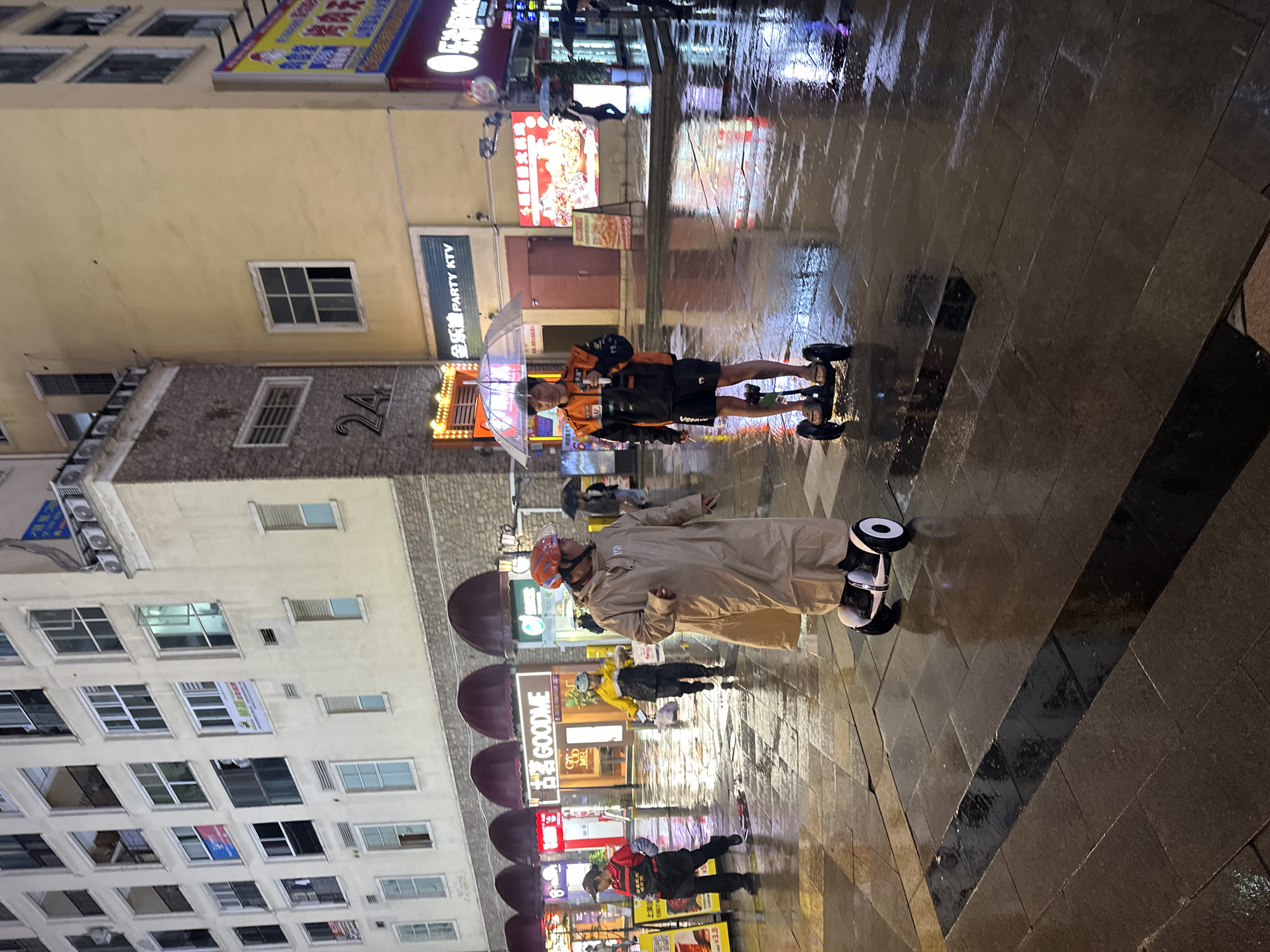 This night-time urban scene captures a bustling street in the Chongqing High-tech Zone, China, on a rainy evening. The paved ground is slick with water, reflecting the vibrant artificial lights from surrounding buildings and signs.
The main subjects are two individuals riding self-balancing scooters (hoverboards) along the wet street. In the foreground, a person wearing a beige raincoat and a helmet with a clear visor navigates on a black and white hoverboard. Further back, another person is seen in an orange and black jacket and black shorts, wearing a mask and holding a clear umbrella, also on a hoverboard. They carry a bag slung over their shoulder. Other people are visible in the background, some holding umbrellas, indicating the persistent rain.
The buildings lining the street are multi-storied with numerous windows and illuminated signs. Notable signs include a vertical one reading "金乐迪 PARTY KTV" (Jin Le Di Party KTV) and another with "古名GOODME" (Gu Ming GOODME) on an awning. The number "24" is prominently displayed on one building, and a small sign shows "¥3.8". Air conditioning units are visible on the exterior walls of some buildings. The overall activity suggests people are commuting or moving about despite the wet weather conditions.