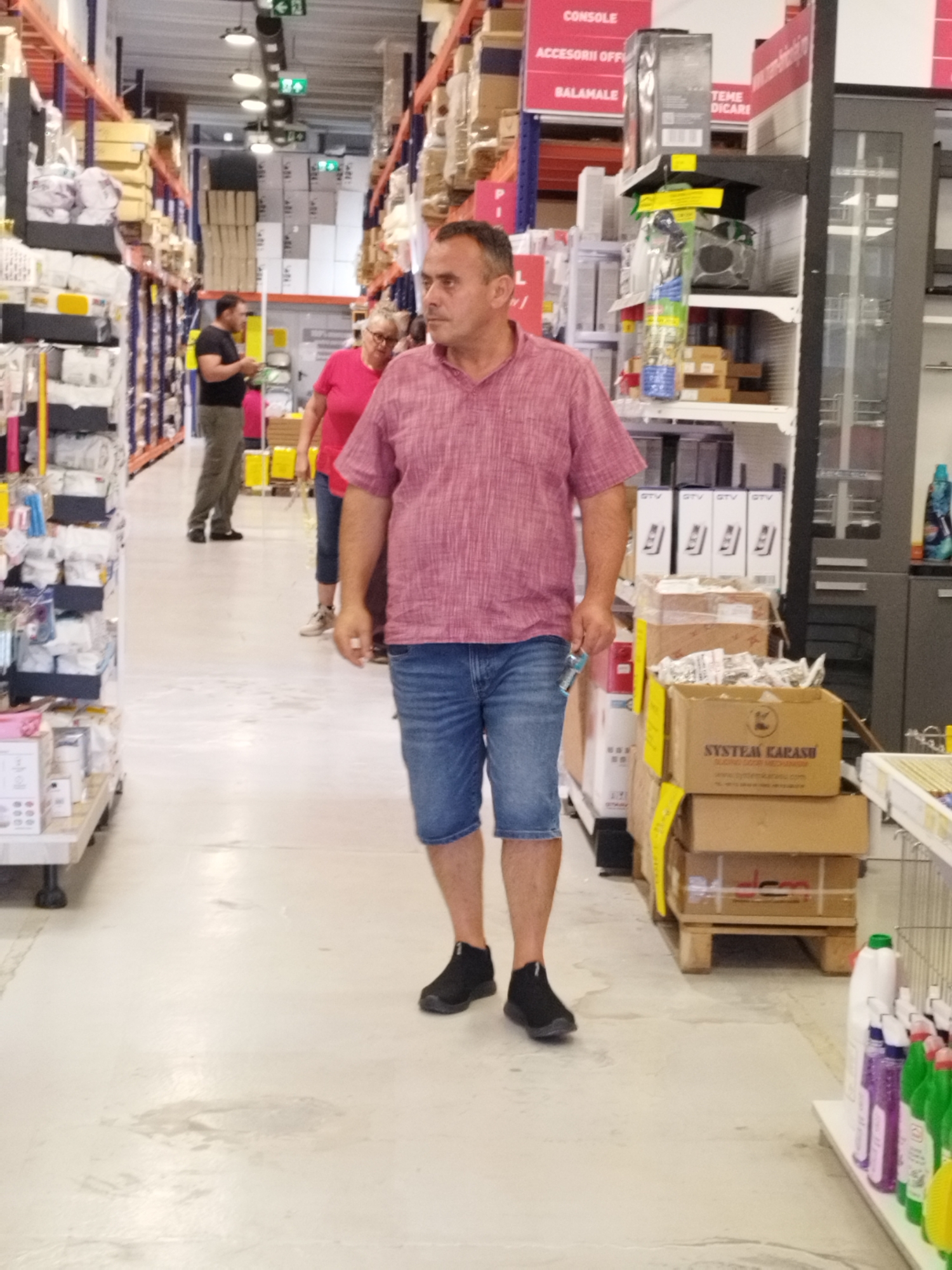 The image shows a man walking down an aisle in a retail store. He is wearing a pink short-sleeved shirt and denim shorts, with black athletic shoes. He is holding a small blue object in his right hand. In the background, other shoppers are visible, including a woman in a pink shirt and a man in a black shirt and grey pants. The store appears to be a large home improvement or electronics store, with shelves stocked with various products. The scene is illuminated by overhead lighting. The store is located in Bucharest, Romania.