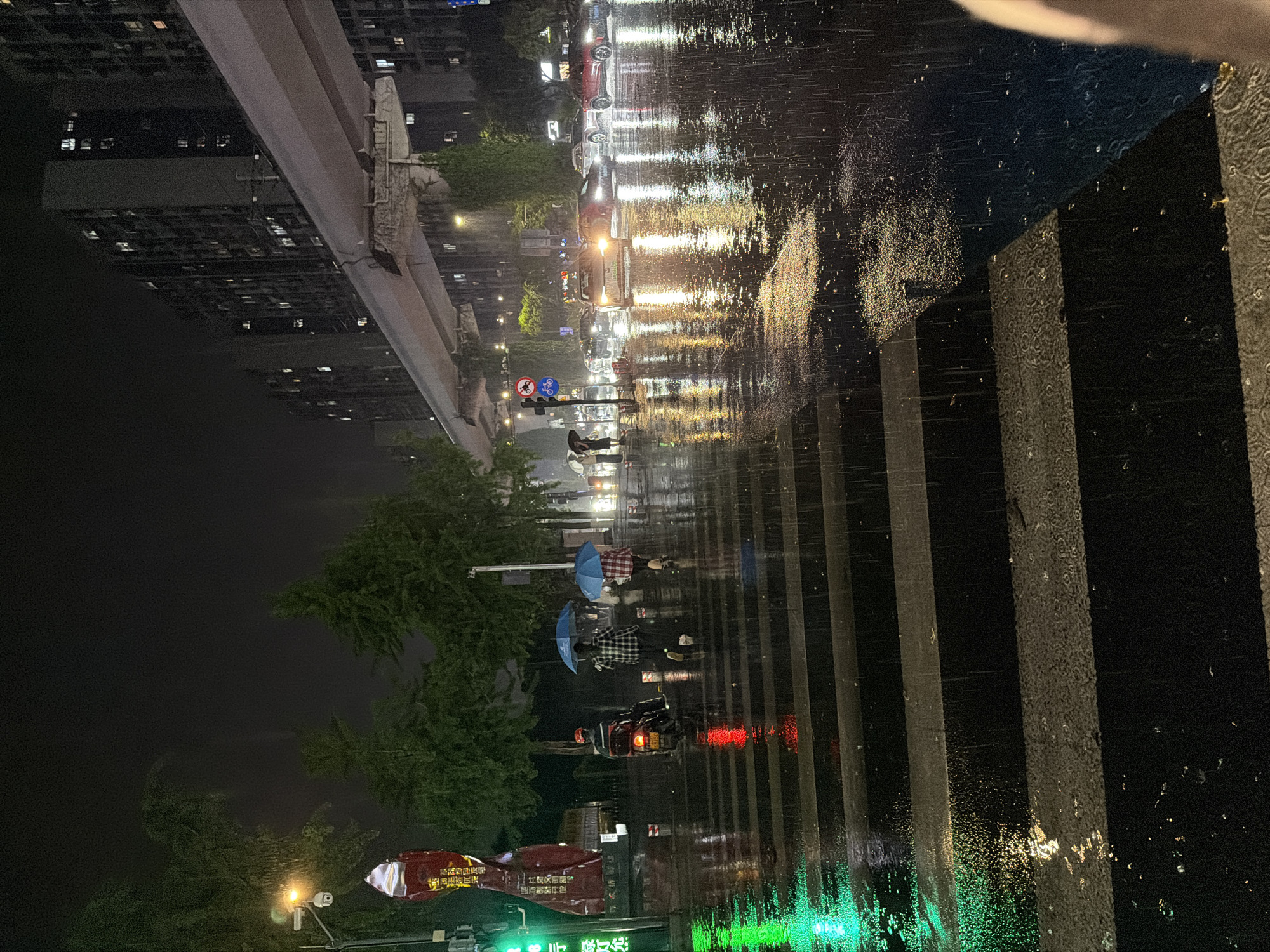 This is a nighttime photograph taken in the Chongqing High-tech Zone, China, showing a street scene in the rain. The camera is positioned near a crosswalk, indicated by white stripes on the wet pavement. People with umbrellas are crossing the street. Several cars and motorcycles with illuminated headlights and taillights are visible on the road. Buildings line the street. Streetlights and neon signs cast reflections on the wet road. Raindrops are visible in the air.