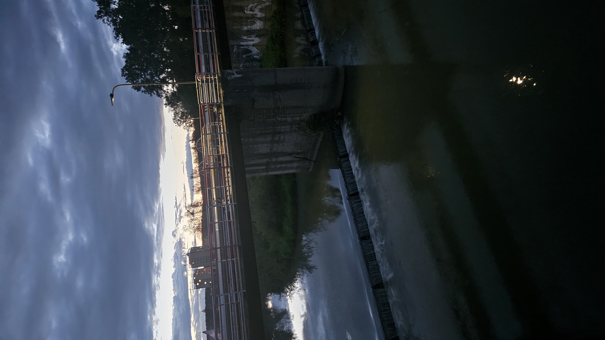 This image depicts a concrete bridge spanning a river with a small waterfall or weir. The sky is overcast with dramatic, dark clouds, suggesting twilight or an approaching storm. A single lamppost is visible on the bridge, illuminated. On the left side of the bridge, buildings can be seen in the distance, possibly part of the city of Prešov, Slovakia. The water in the river is calm, with reflections of the sky and bridge. Trees and vegetation line the banks of the river. The scene is peaceful and slightly moody.