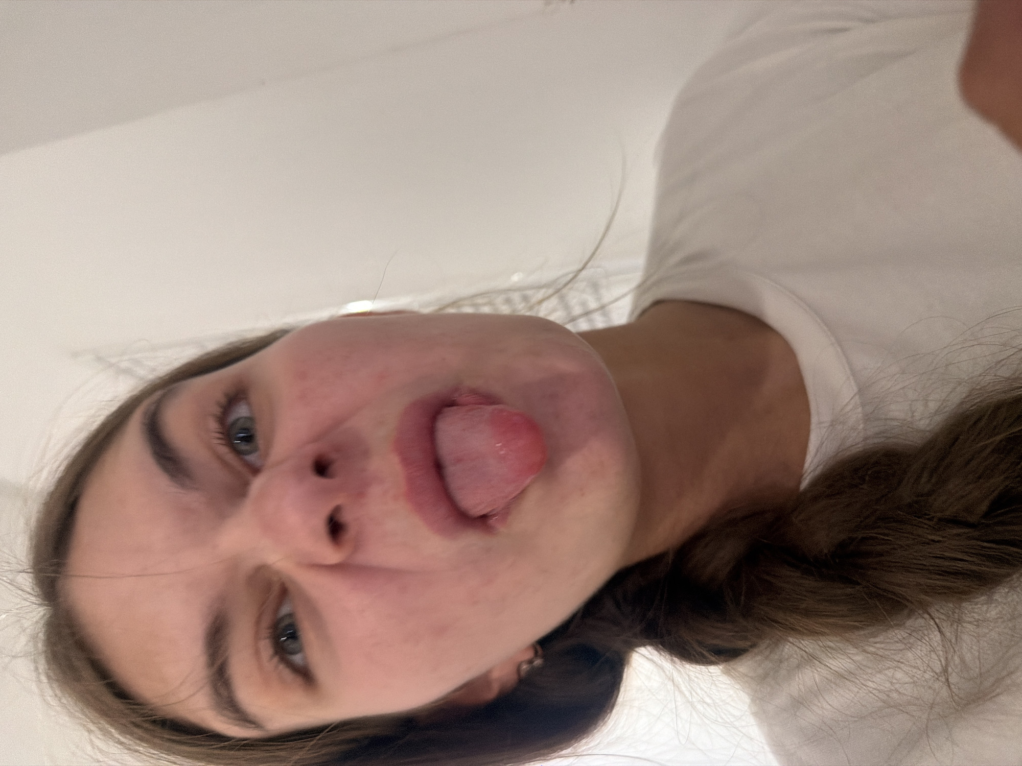 A young woman with brown hair in a braid is sticking her tongue out at the camera. She is wearing a white t-shirt. The background is a plain, light-colored wall, possibly indoors. There are no specific visual cues in the image that would indicate a location in Nha Trang, Vietnam.
