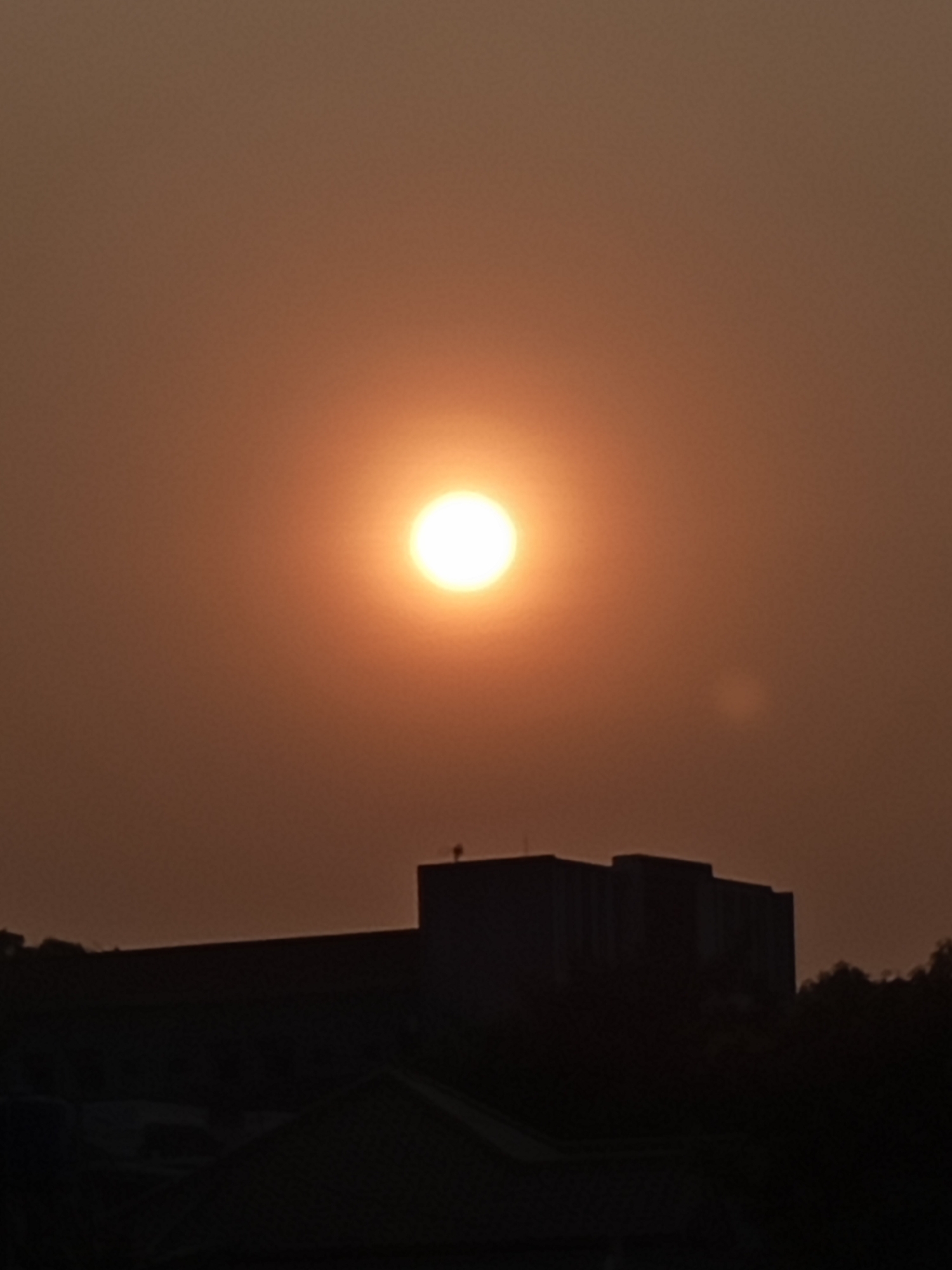 A vertical, medium shot captures the sunset over Semarang, Indonesia. The sun, a bright, glowing orb, is positioned slightly left of the center of the frame, casting a warm, orange light that suffuses the hazy sky. Below the sun, silhouetted against the luminous sky, is the dark outline of a building, appearing to be a multi-story structure. The building's geometric form suggests a modern architectural style. The foreground is dominated by the dark shapes of lower buildings and trees, also rendered as silhouettes, indicating they are significantly darker than the illuminated sky. The overall impression is one of a city at dusk, with the sun setting amidst atmospheric haze.