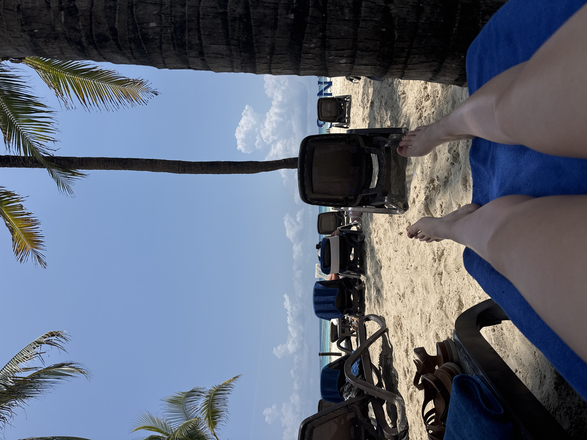 The image depicts a beach scene, likely taken in Playa del Carmen, Mexico, although there is nothing in the image to positively ID the location. The view is from the perspective of someone relaxing on a blue towel-covered lounge chair, with their legs and feet visible in the foreground. The background shows a row of empty lounge chairs on the sandy beach. The sea is visible in the distance. Two dark tree trunks frame the scene, with palm fronds visible near the edges of the image. The sky is predominantly blue with scattered clouds.