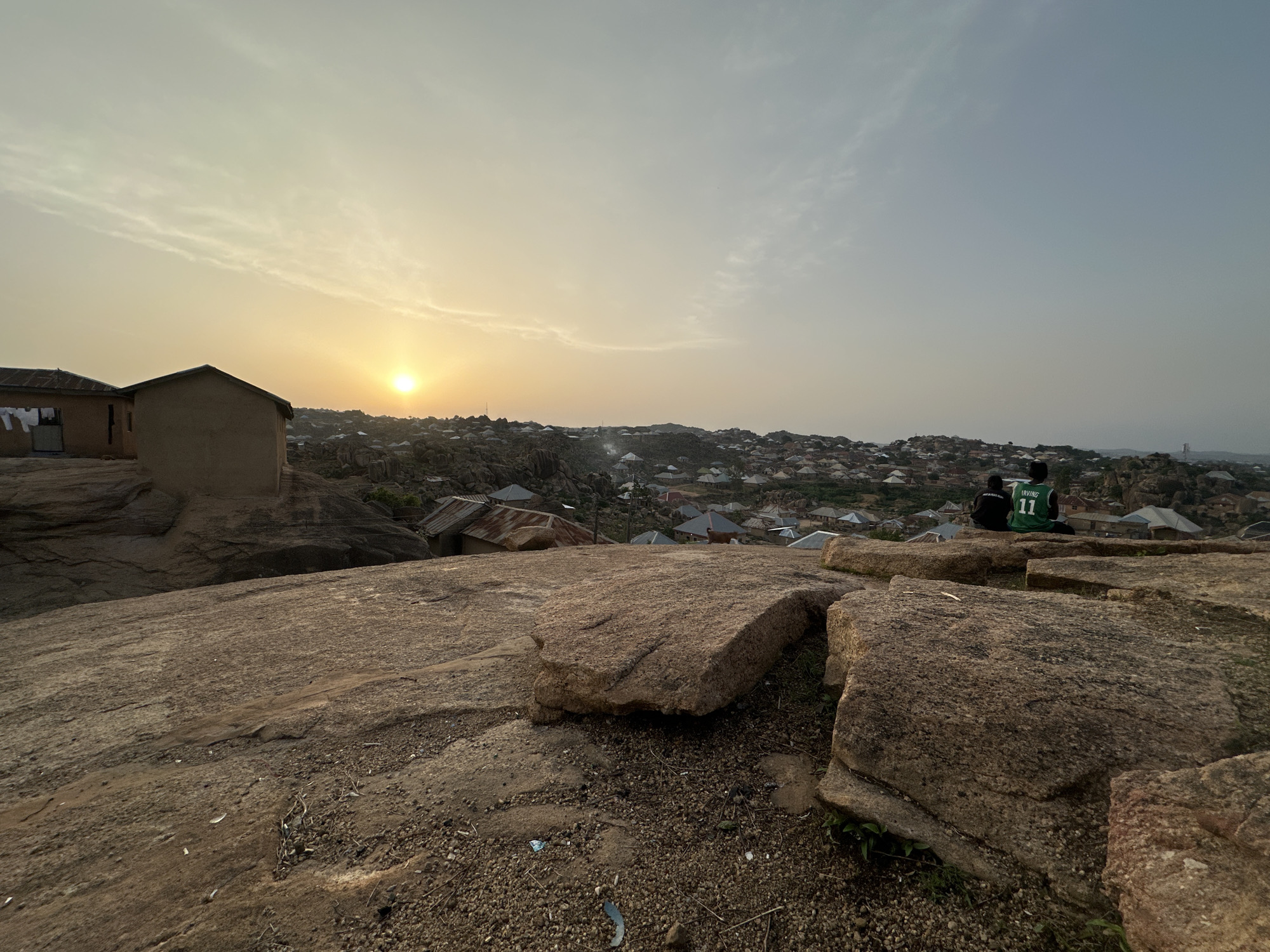 Jos, Nigeria. A wide angle shot taken at ground level shows the summit of a large, rocky hill. Two people sit overlooking a dense residential area and the setting sun. The foreground is composed of a wide, flat rock covered with small loose gravel. A crude brick structure stands to the left of the frame with clothes hanging on a line outside. The sun is low on the horizon in the center of the frame, partially obscured by a haze. The sky is pale yellow and orange near the sun and transitions to a light blue gray at the top of the frame. The residential area is comprised of numerous small buildings with metal roofs. Two individuals are seated together in the midground on a large boulder, facing away from the camera. The person on the right wears a green and black basketball jersey with the name "IRVING" and the number "11" visible on the back. The person on the left wears a black jacket or shirt.