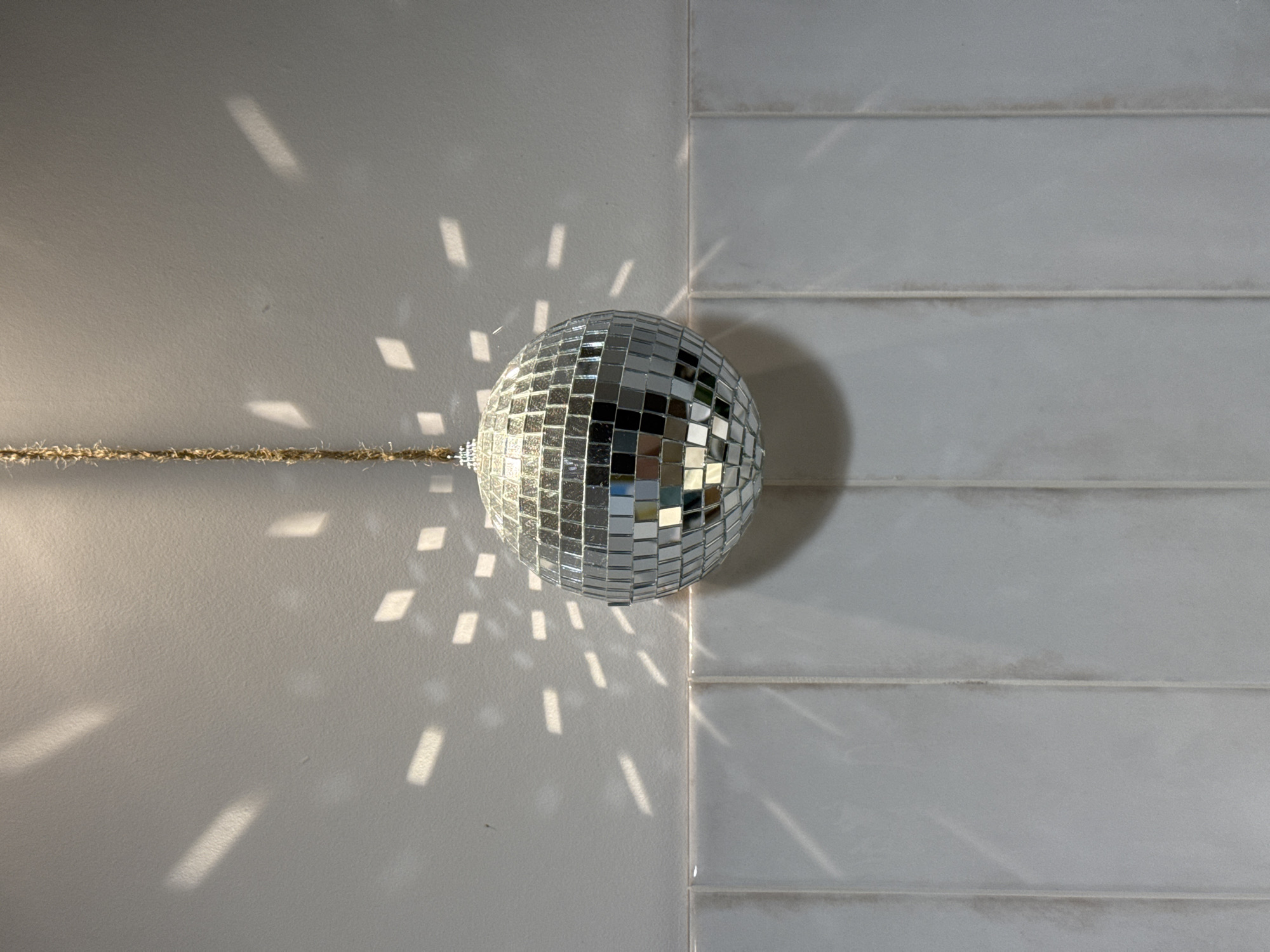 A disco ball hangs from a thin brown string against a wall composed of two different textures. To the left is a smooth, solid surface while the right side features horizontally-laid rectangular tiles with visible grout lines. Light reflects off the disco ball, creating scattered bright spots and streaks on both sections of the wall.