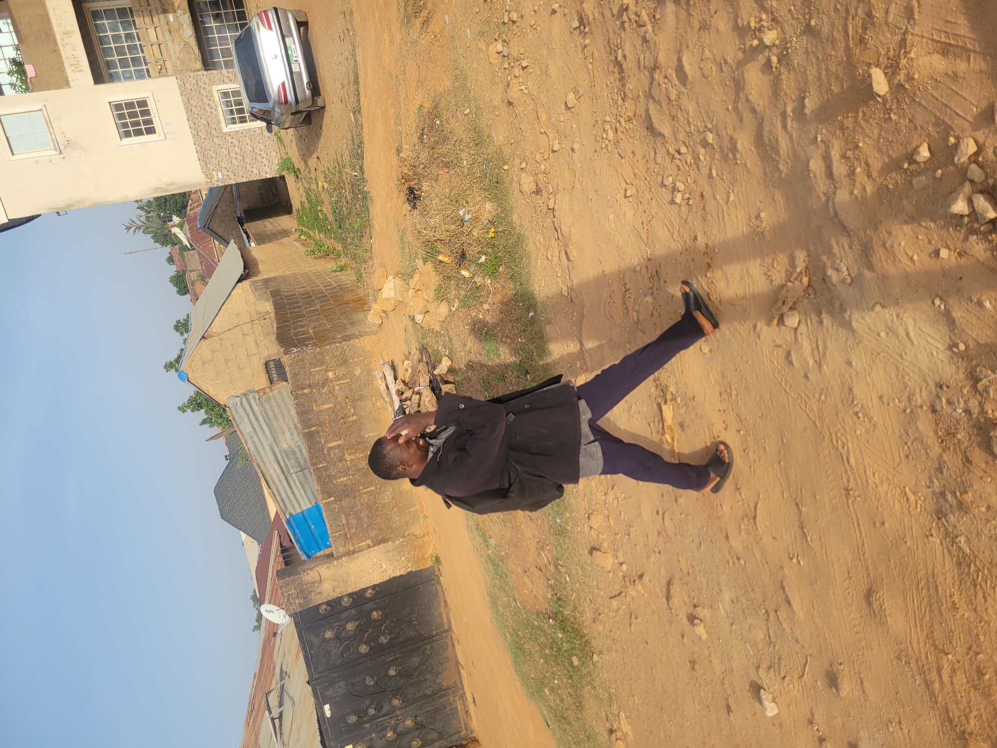 The image captures a young man standing on a dusty, unpaved ground in what appears to be a residential area in Jos, Nigeria. The man, positioned centrally, is wearing a dark jacket over a lighter shirt, purple trousers, and dark sandals. His left arm is raised with his hand near his face, possibly shielding his eyes from the bright sun or in a casual gesture, while his legs are spread slightly apart.
In the background, there are several buildings, predominantly constructed from concrete. A two-story building with multiple windows, some featuring white frames or security bars, is visible, and its lower section exhibits a patterned stone or brick facade. A silver sedan, likely a Peugeot, is parked next to this building, displaying a green and white license plate, though the text is unreadable. Further back, other structures with corrugated metal roofs are visible, and a decorative dark metal gate can be seen on the left. The sky is clear and bright blue.
The ground beneath the man's feet is primarily dry, sandy dirt with scattered small rocks and sparse patches of dry grass and weeds. Some minor litter is visible on the ground. Long shadows are cast, indicating bright sunlight and suggesting a time of day like morning or late afternoon. The scene depicts a typical outdoor setting in a developing urban or suburban environment, consistent with the provided location context of Jos, Nigeria. No discernible text is clearly legible other than the general format of the car's license plate.
