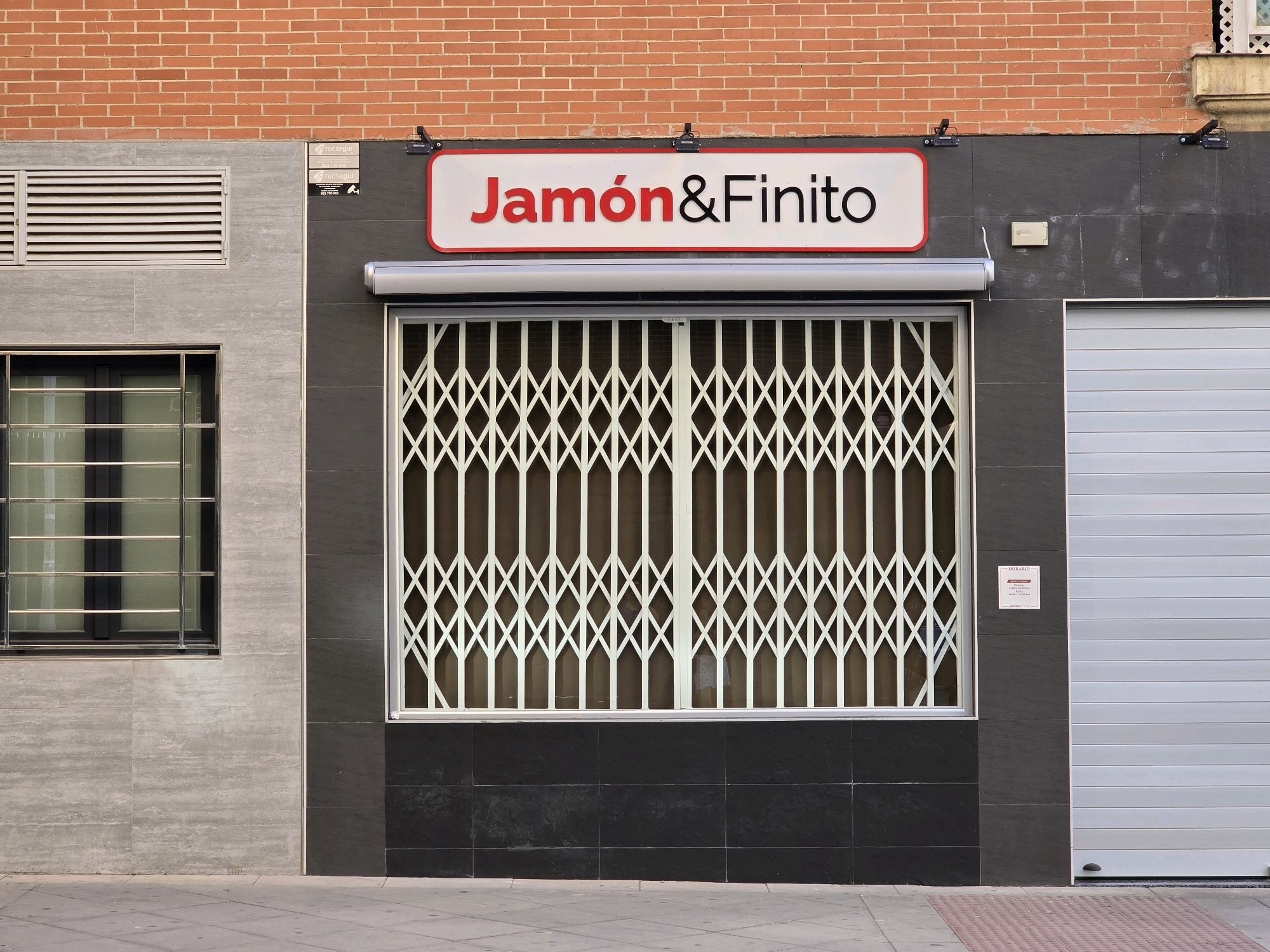 The image shows the exterior of a storefront in Jaén, Spain. The storefront features a red brick facade on the upper portion and a dark tiled facade below. A large, white sign with red and black lettering reads "Jamón&Finito". Below the sign, a large window is protected by a white, expandable metal grate. To the left of this window, there is another, smaller window with metal bars. To the right, a light gray, corrugated metal garage door is visible. The sidewalk in front of the storefront is paved with gray concrete tiles and a red textured tactile paving. The lighting suggests it is daytime, with sunlight casting subtle shadows. The scene appears to be a commercial establishment, likely a shop or restaurant.