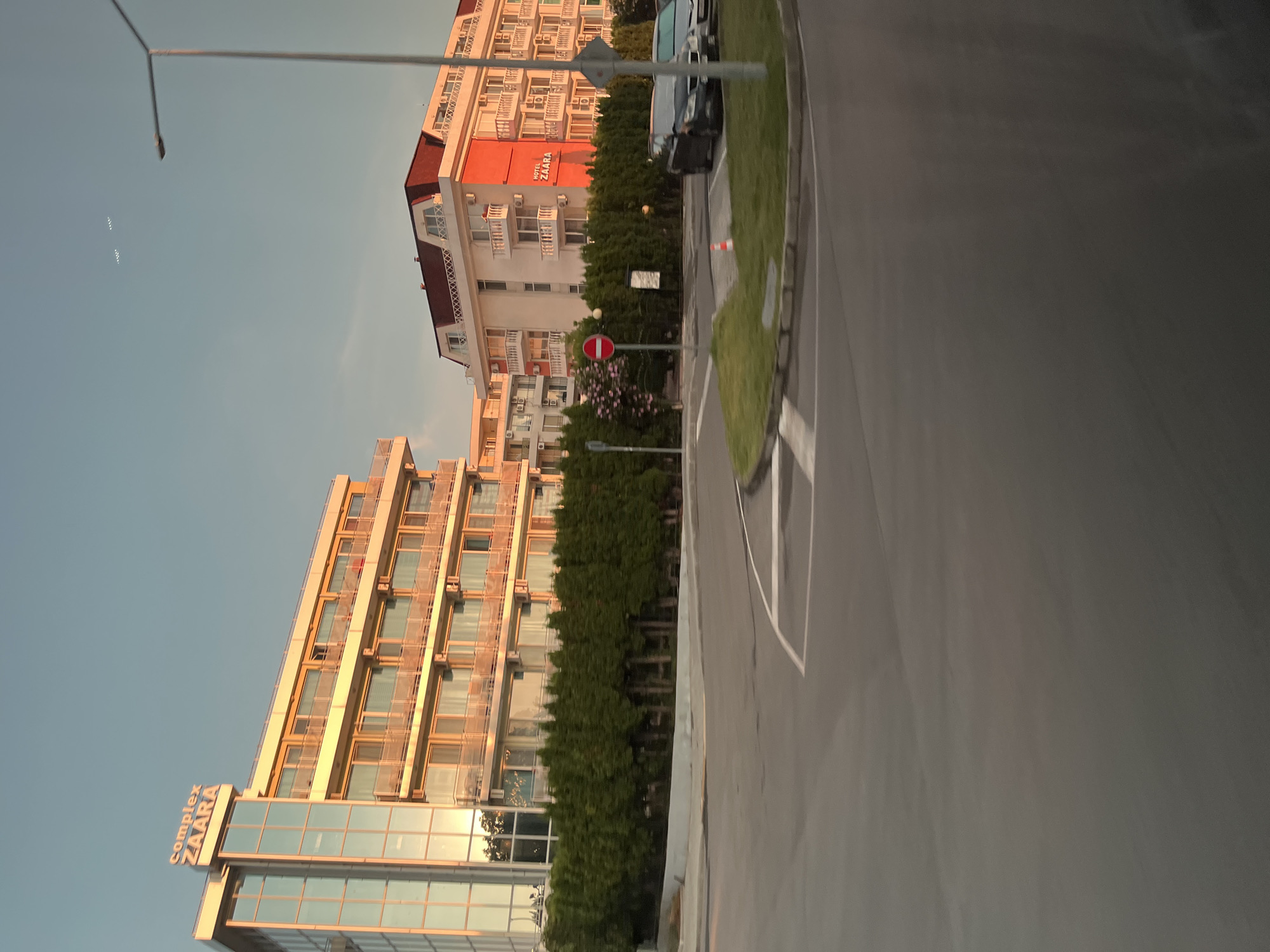 The image captures a scene in the city of Nesebar, Bulgaria, likely during the late afternoon or early evening, given the warm, golden light illuminating the buildings. In the foreground, a paved road curves to the left, with white lane markings visible. A well-maintained green lawn with a small orange traffic cone is situated on the inner curve of the road.
Dominating the background are two multi-story buildings. The building on the left is taller, with a modern glass facade and balconies. The words "Complex ZAARA" are prominently displayed in white lettering at the top. The building on the right is also multi-story, with a distinctive red-tiled, mansard roof. One section of this building features a bright orange facade, and the words "HOTEL ZAARA" are visible in red lettering. Both buildings appear to be hotels or complexes of apartments, characterized by numerous balconies and windows. A line of dense, dark green trees forms a visual barrier between the buildings and the road.
Several vehicles are parked on the right side of the image, including a dark-colored sedan and a lighter-colored car behind it. A street lamp stands tall on the right side of the frame, extending into the clear, pale blue sky. In the sky, two small, bright, geometric shapes are visible, possibly distant aircraft or drones. There is no visible human presence in the image. The overall impression is one of a modern resort or residential area bathed in pleasant evening light.