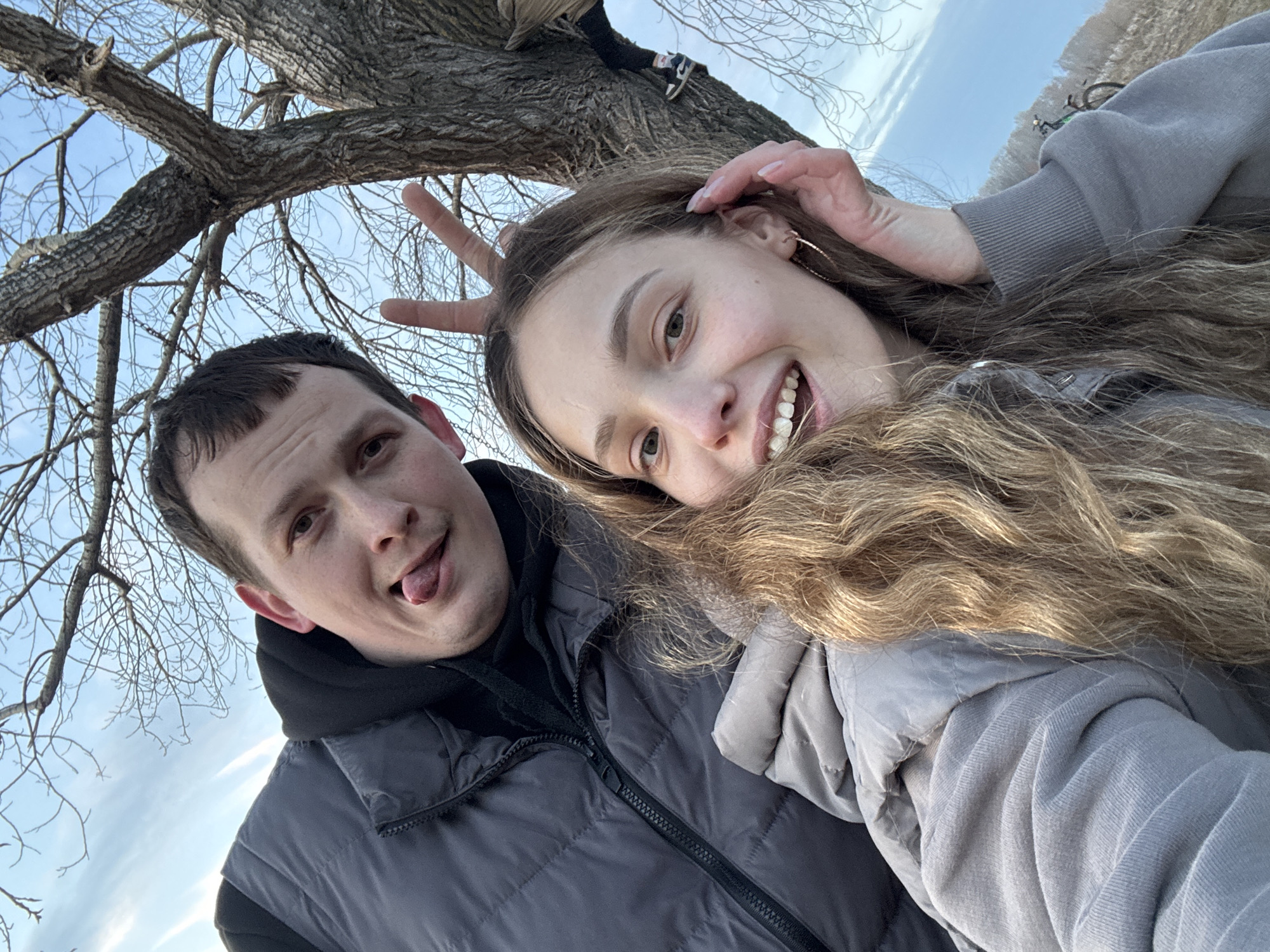 A low-angle selfie photograph features two individuals under a tree. A man on the left with short dark hair and wearing a gray vest and black hooded shirt, sticks out his tongue. A woman on the right, wearing a gray sweatshirt, holds her fingers up behind her head in a "bunny ears" gesture. The tree has bare branches, and a person is sitting in the tree above. The sky is visible in the background. The picture was taken in the city of Chaikovsky, Russia.