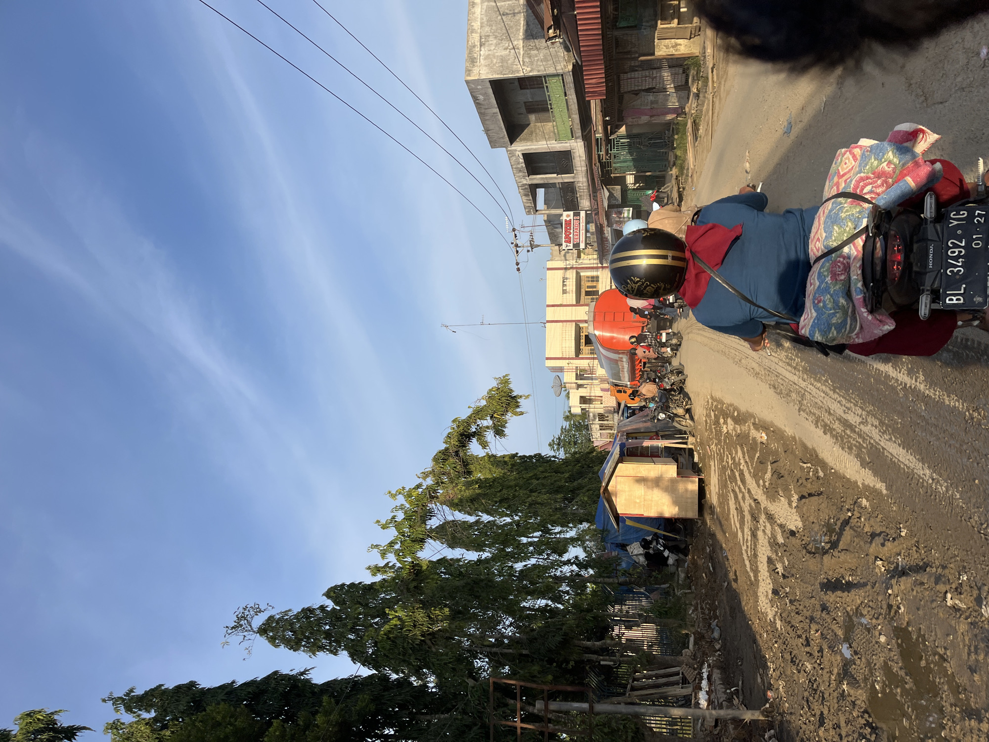 The image depicts a street scene in Alurcucur, Indonesia, characterized by a muddy, unpaved road. A person is riding a motorcycle in the foreground, from behind, wearing a blue shirt, red pants, and a black and gold helmet. A red scarf is draped around their neck, and a colorful blanket is secured to the back of the motorcycle. Other motorcycles and a large orange tanker truck are visible further down the road, along with several buildings lining the street. The sky is clear and blue with some wispy clouds. Vegetation, including tall trees and smaller plants, is present on the left side of the road. The overall impression is one of a developing or rural area with basic infrastructure.