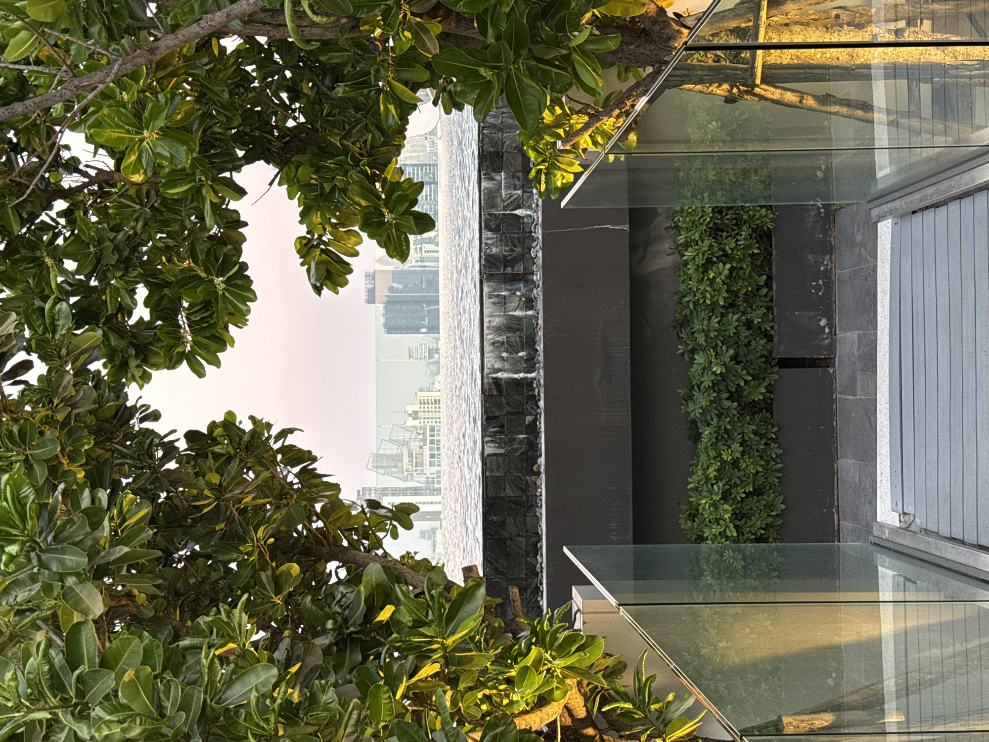 The image is shot from an elevated position in Bangkok, Thailand, overlooking the city skyline. A tree with lush green leaves occupies the left and upper parts of the frame. A modern structure with glass panels and grey stonework is visible on the right. A cascading water feature with a textured stone wall separates the structure from the city view. The water feature reflects the urban landscape, creating a mirror-like effect. A vertical row of green plants is situated near the glass panels. The sky is a pale, muted color, indicating either dawn or dusk. The scene conveys a blend of nature and modern architecture.