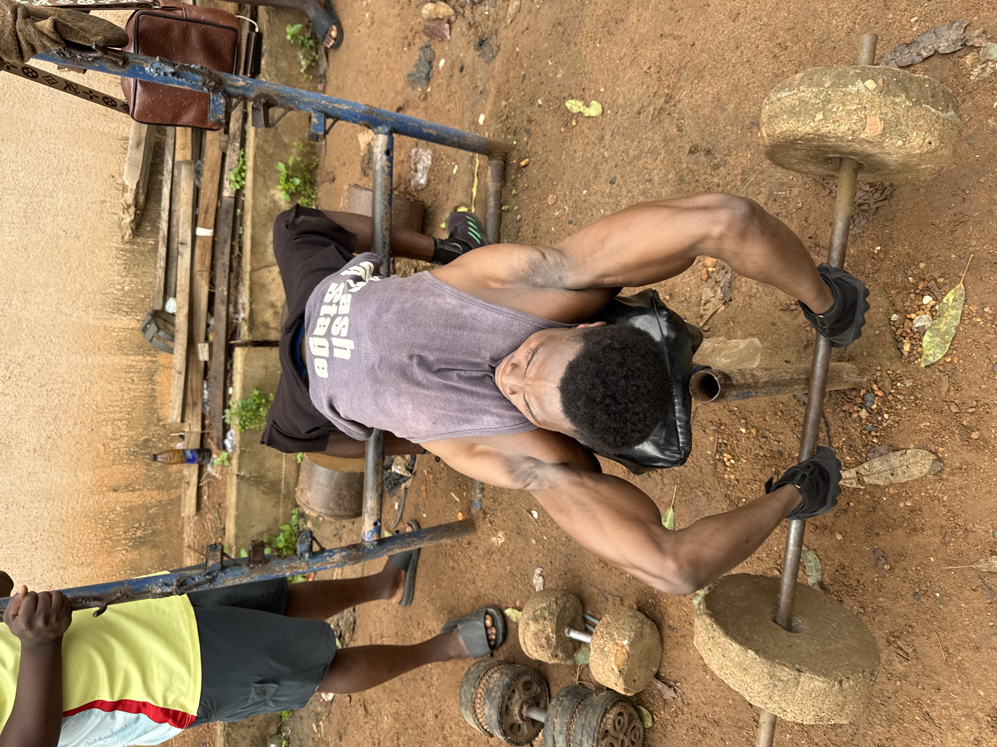 A person is exercising with weights outdoors. The scene is set on dirt ground with some scattered leaves and small stones, suggesting an unpaved outdoor area. A metal frame, possibly part of a weightlifting apparatus, is visible in the background.
The main subject is a person, seen from a top-down perspective, lying down and lifting a barbell. They are wearing a grey tank top with text that is partially visible and appears to say "Wash Stage". Black weightlifting gloves are on their hands, and they are also wearing dark shorts. Two weights, made of what appears to be stone, are attached to the barbell. Nearby, on the ground, are additional homemade weights, some resembling old car parts with exposed gears.
Another person is partially visible on the left side of the frame, wearing a yellow t-shirt and dark shorts. They are standing and appear to be holding onto the metal frame.
The lighting suggests it is daytime, with no strong shadows indicating early morning or late afternoon. The environment appears somewhat rustic, possibly a makeshift gym. There are no clear indicators of the weather, but the dry dirt suggests it might not be raining. The overall mood is one of focused physical exertion.