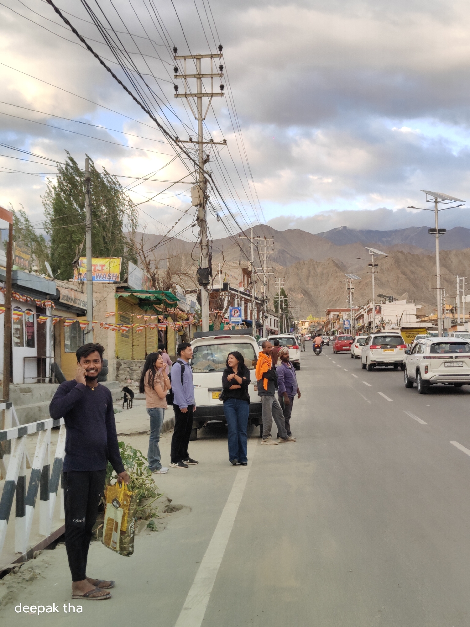 The image depicts a street scene in the city of Leh, India, with a backdrop of majestic mountains under a cloudy sky. The street is lined with shops and buildings, one of which displays a sign for "TT Wash". A prominent electrical pole with numerous wires crisscrosses the sky, indicating the presence of urban infrastructure. Several people are gathered on the side of the road, some appearing to be waiting for transportation or engaging in conversation. Among them, a man in a dark blue long-sleeved shirt and black pants is holding a large shopping bag and talking on his phone. A white car is parked nearby, and other vehicles are visible on the road, suggesting moderate traffic. Prayer flags are strung across some of the buildings, adding a cultural element to the urban landscape. The ground appears to be dusty, typical of arid regions.