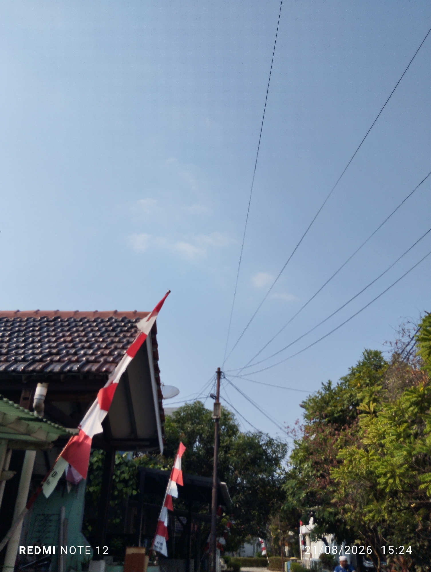 A low-angle, outdoor, daytime photograph captures a residential street in Bekasi, Indonesia, under a clear blue sky. Several utility wires are strung across the upper portion of the frame, converging towards the right. In the foreground, on the left, a portion of a house is visible, featuring a dark tiled roof and a white downspout. A red and white Indonesian flag is mounted on the side of the house, angled upwards. Further down the street, another Indonesian flag is visible, along with lush green trees and foliage. The bottom edge of the image displays a timestamp and device information: "21/08/2026 15:24" and "REDMI NOTE 12". No people are clearly identifiable in the scene. The overall impression is one of a quiet, sunny day in a suburban neighborhood.