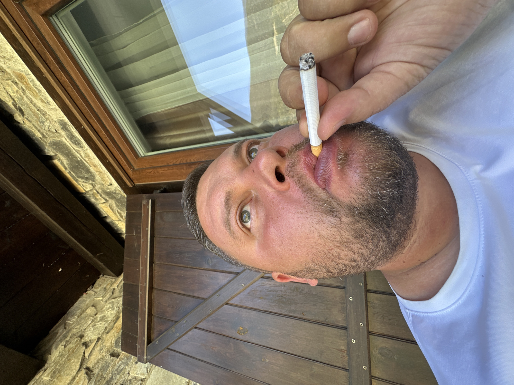 The image features a man actively smoking a cigarette, seen in a close-up, rotated perspective. The man, appearing to be in his 30s to 50s, has short dark hair and a salt-and-pepper beard. He wears a plain white t-shirt and looks directly towards the camera with light-colored eyes, holding a lit cigarette with a visible ash tip to his lips.
The setting appears to be a rustic building, likely in Encamp, Andorra. To the left of the man, a rough-hewn stone wall is visible, suggesting traditional construction. Above and behind him, a wooden-framed window or skylight is present, through which internal blinds or curtains can be seen, along with reflections of a bright sky and possibly white clouds or structures. To the right and below the man, dark wooden panels or shutters, fastened with visible nails, form part of the background, along with dark wooden beams.
The lighting is bright and natural, indicating it is daytime. No visible text is discernible on the man's clothing, the cigarette, or the surrounding elements. The man's expression is neutral, possibly inquisitive, as he gazes straight ahead while smoking.