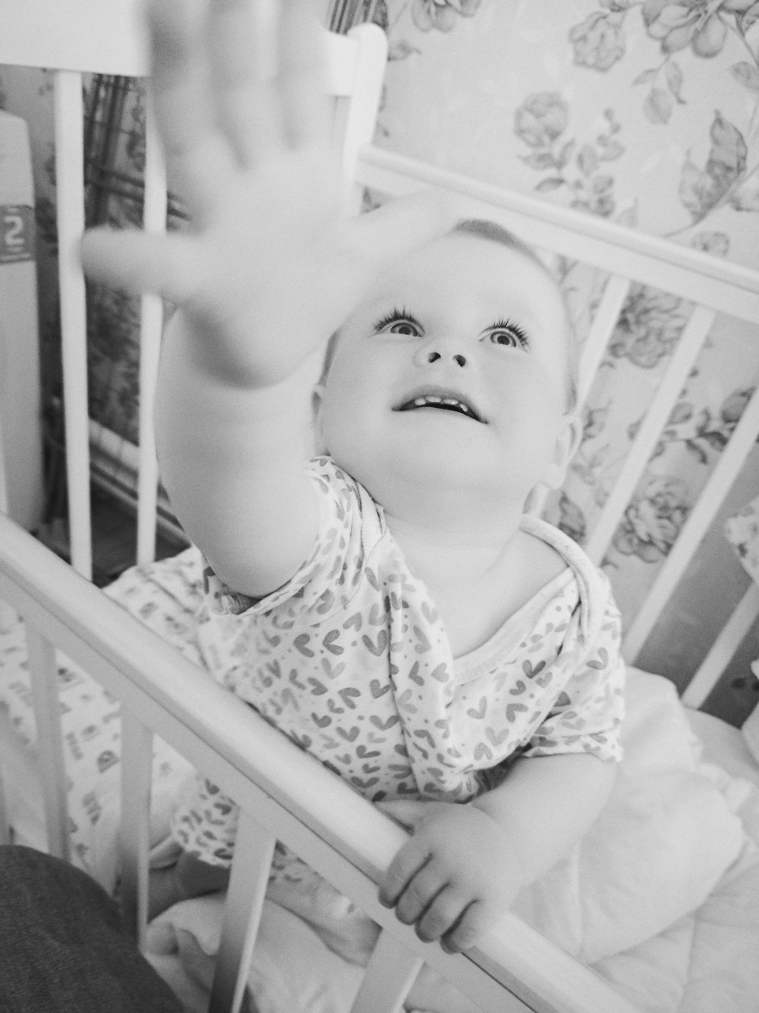 A baby is in a crib in a room with floral wallpaper. The baby has one arm outstretched, reaching upwards. The location is unspecified except that it is in Фоки, Russia.