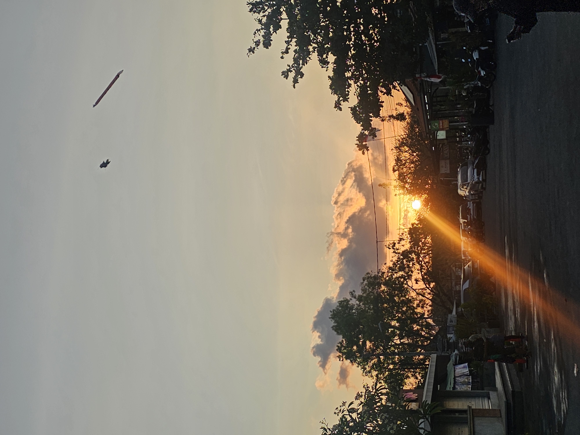 A vertical, medium shot captures a sunset over a parking area adjacent to shops in Denpasar, Indonesia. The sun, a bright orb, sits low on the horizon, its rays casting a strong, orange lens flare diagonally across the scene from the bottom left upwards. The sky is a gradient of pale yellow and soft orange, with scattered clouds tinged with pink and grey. In the sky, two objects are visible: a small, dark bird, possibly a kite, and a long, red, cylindrical object, also appearing to be a kite or banner.
In the foreground, a dark asphalt surface is visible, reflecting some of the sunset's light. A few parked cars and motorcycles are scattered across the area. To the left, a small, single-story building with a gray roof and light-colored walls has laundry hanging on display outside. Two figures, appearing to be children, stand near the building. Further back, more trees and buildings are silhouetted against the sunset.
To the right, the dense foliage of trees obscures the view. A person wearing a dark jacket and a baseball cap is partially visible on the far right edge, looking down at their phone. The overall impression is a serene, end-of-day scene in a moderately busy urban setting.