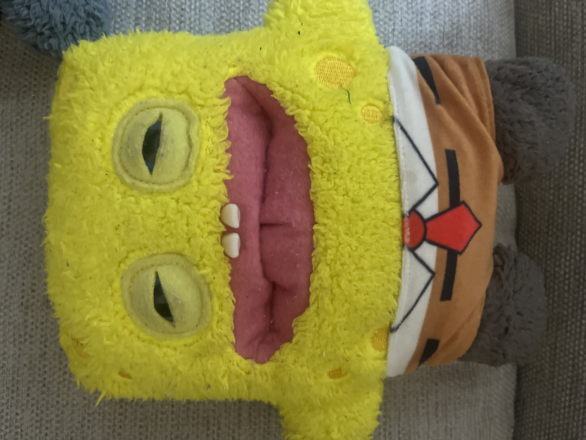 This is a close-up, eye-level, waist-up shot of a stuffed Spongebob Squarepants toy. The toy is yellow and fuzzy, with large, half-closed eyes and a wide, open mouth revealing pink interior and white teeth. It is wearing a brown shirt with a white collar and a red tie. The toy is resting on a gray, textured surface, possibly a couch. There are no visible indications of the city of Aktau, Kazakhstan, in the image.