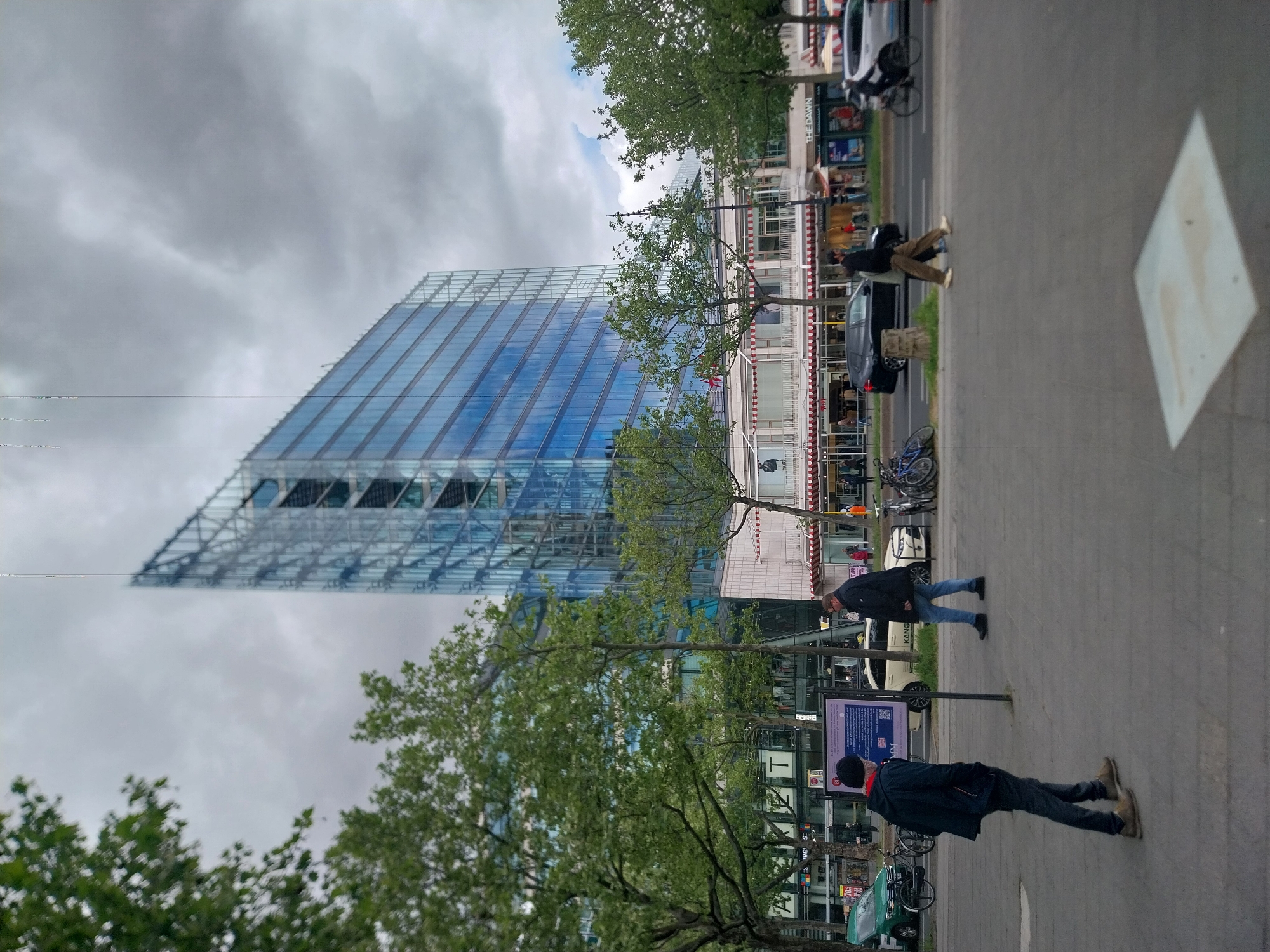 The image captures a street scene in Berlin, Germany, featuring the striking high-rise building, the Sony Center. The modern architectural structure is characterized by its glass facade, visible against a cloudy sky. The foreground shows people standing and walking on a sidewalk adjacent to the street. A sign post with informational text and QR code stands prominently. Cars and bicycles are parked on the roadside. The surroundings include buildings and trees, adding to the urban landscape.
