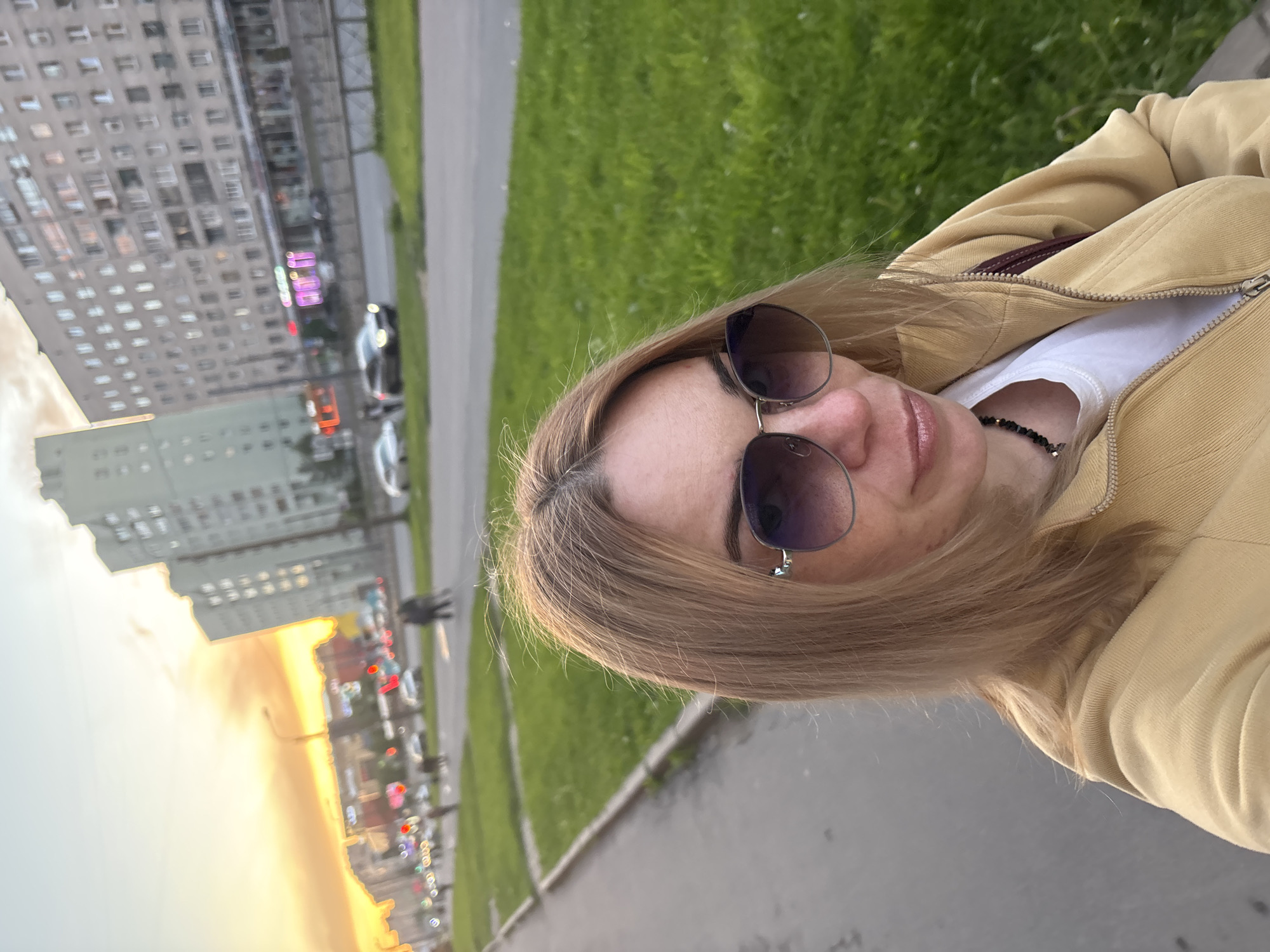 In Sinyavino, Russia, a woman with blonde hair and sunglasses is taking a selfie on a sidewalk. She is wearing a yellow hooded jacket over a white t-shirt and a beaded necklace. Behind her, a grassy area borders a street with cars and multi-story buildings. The sky is filled with warm hues of orange and yellow, indicating either sunrise or sunset.