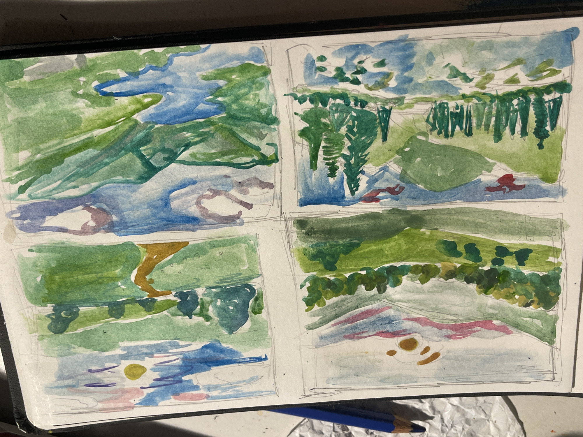 The image shows a sketchbook page with four roughly painted landscape sketches using watercolor. Each sketch is contained within a roughly drawn square frame. The sketches appear to depict generalized scenes of water, trees, and hills. The top left sketch contains a blue river or stream flowing through green land. The top right shows a forest of thin trees in dark green, with foliage overhead and a blue sky above. The lower left sketch depicts a green field with a brown pathway bisecting it. The bottom right sketch features a hill with a line of trees at its base, with a pink and blue water scene in the foreground. The sketches are rudimentary and impressionistic. The sketchbook sits on a surface. A blue colored pencil and crumpled silver foil are visible near the bottom of the image. The sketches do not appear to relate to a location in Maidstone, United Kingdom.