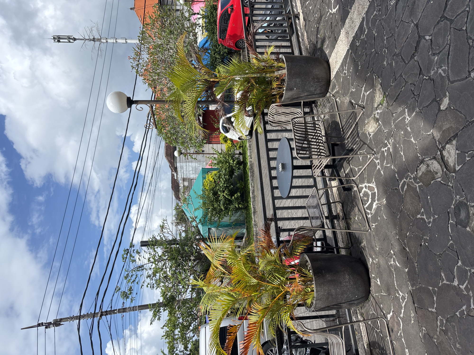 The scene is an outdoor seating area in Kuta Selatan, Indonesia. Two large, dark gray planters contain palm trees with green and yellow fronds. Between the planters is a metal table with two matching chairs. The ground is paved with dark gray stones with white grout. Behind the seating area is a low metal fence. Beyond the fence, there are parked vehicles, including a red car and a white van, and lush greenery. Buildings of varying heights and colors are visible in the background. Overhead, power lines stretch across a partly cloudy sky. A white globe streetlight and a telecommunications tower are also visible.