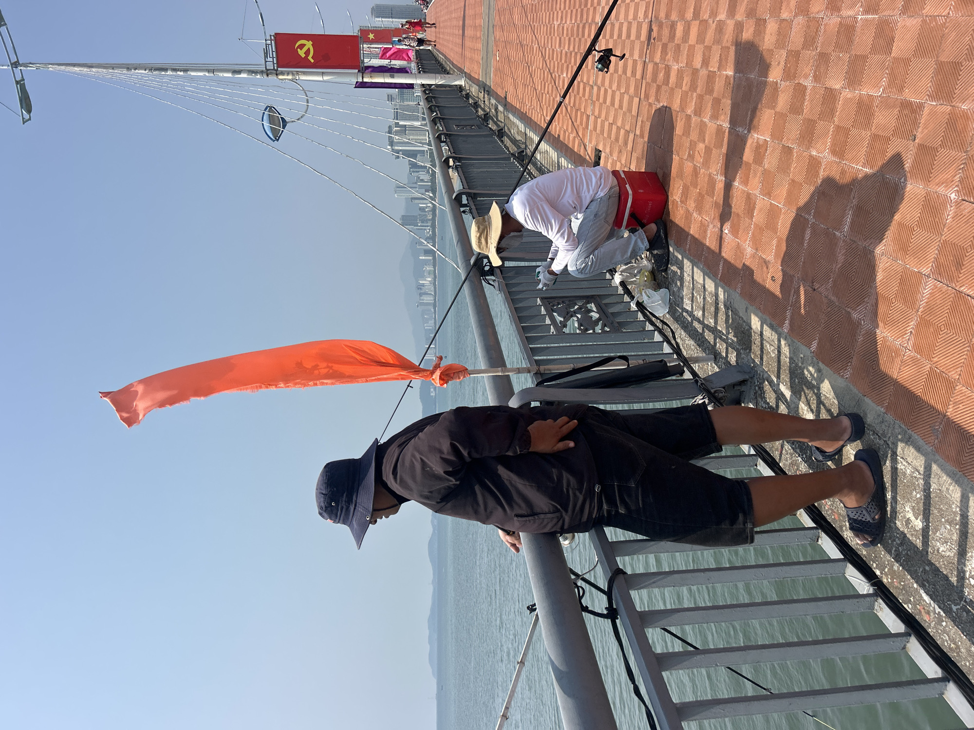 A wide-angle, outdoor shot depicts two people fishing on a bridge or pier, potentially in or near Bắc Nha Trang, Vietnam, indicated by the Chinese-style communist flag visible in the background.
One man, wearing a dark jacket, dark shorts, a navy blue hat, and sandals, leans against the railing, looking down at the water. An orange flag is tied to the railing near him.
Another person, wearing a white long-sleeved shirt, light grey pants, a tan straw hat, and dark-colored sandals, kneels on a red bucket. This person seems to be preparing fishing gear or bait. Fishing rods are positioned nearby.
The bridge or pier has a red, patterned tile surface and a metal railing. The sea is visible below. In the background, there are buildings and other structures that may be part of the city skyline. The sky is clear and blue.