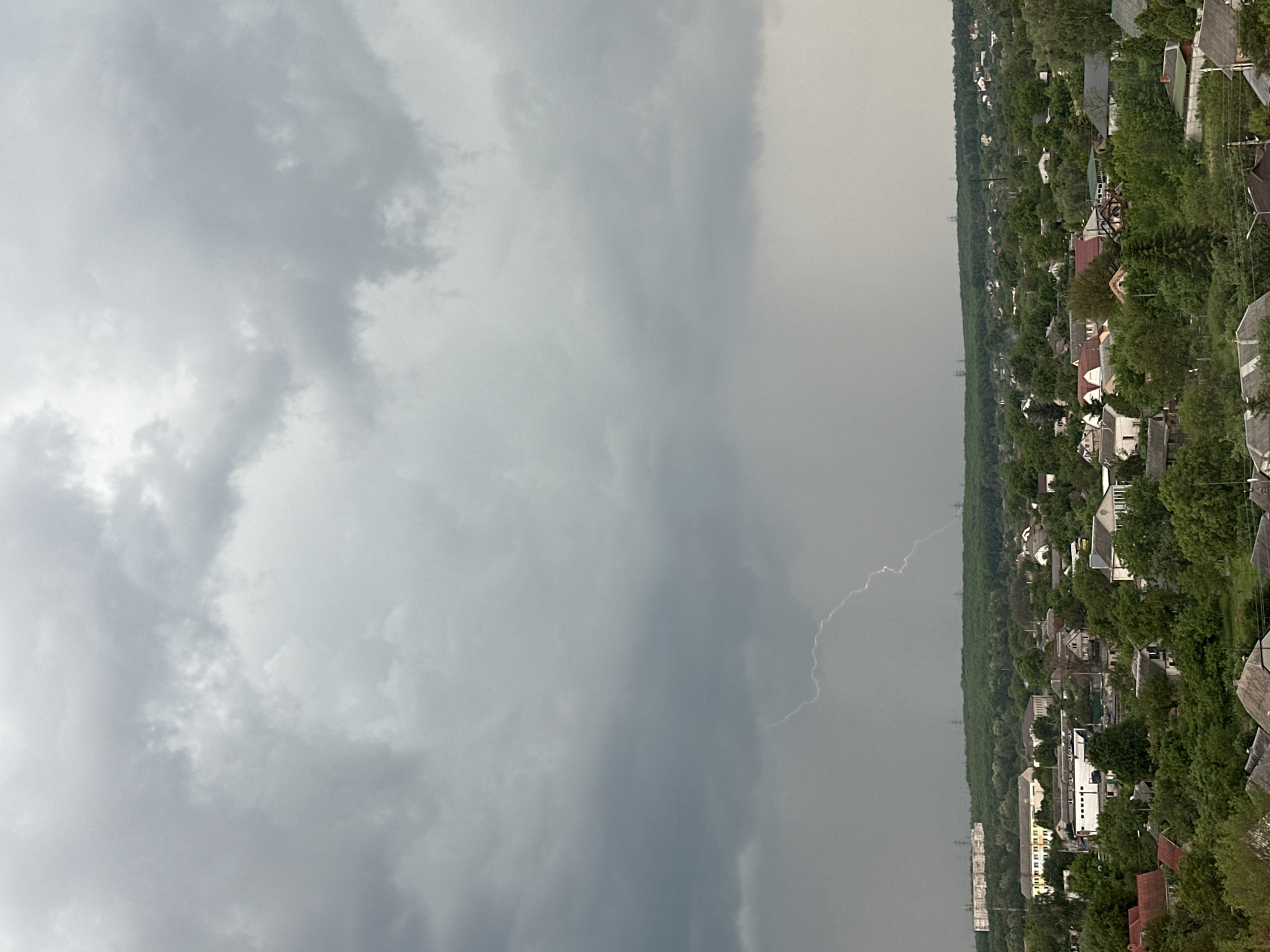 The image shows a landscape of a city under a stormy sky, likely taken from an elevated perspective. The sky is dominated by dark, ominous clouds, with a lightning strike visible in the center, illuminating the scene. Below the sky, a dense cityscape stretches out, composed of buildings, trees, and infrastructure. The buildings appear to be a mix of residential and commercial structures. The density of trees suggests a suburban or green area within the city. The lightning strike serves as a focal point, emphasizing the intensity of the storm. The scene conveys a sense of foreboding and the power of nature.