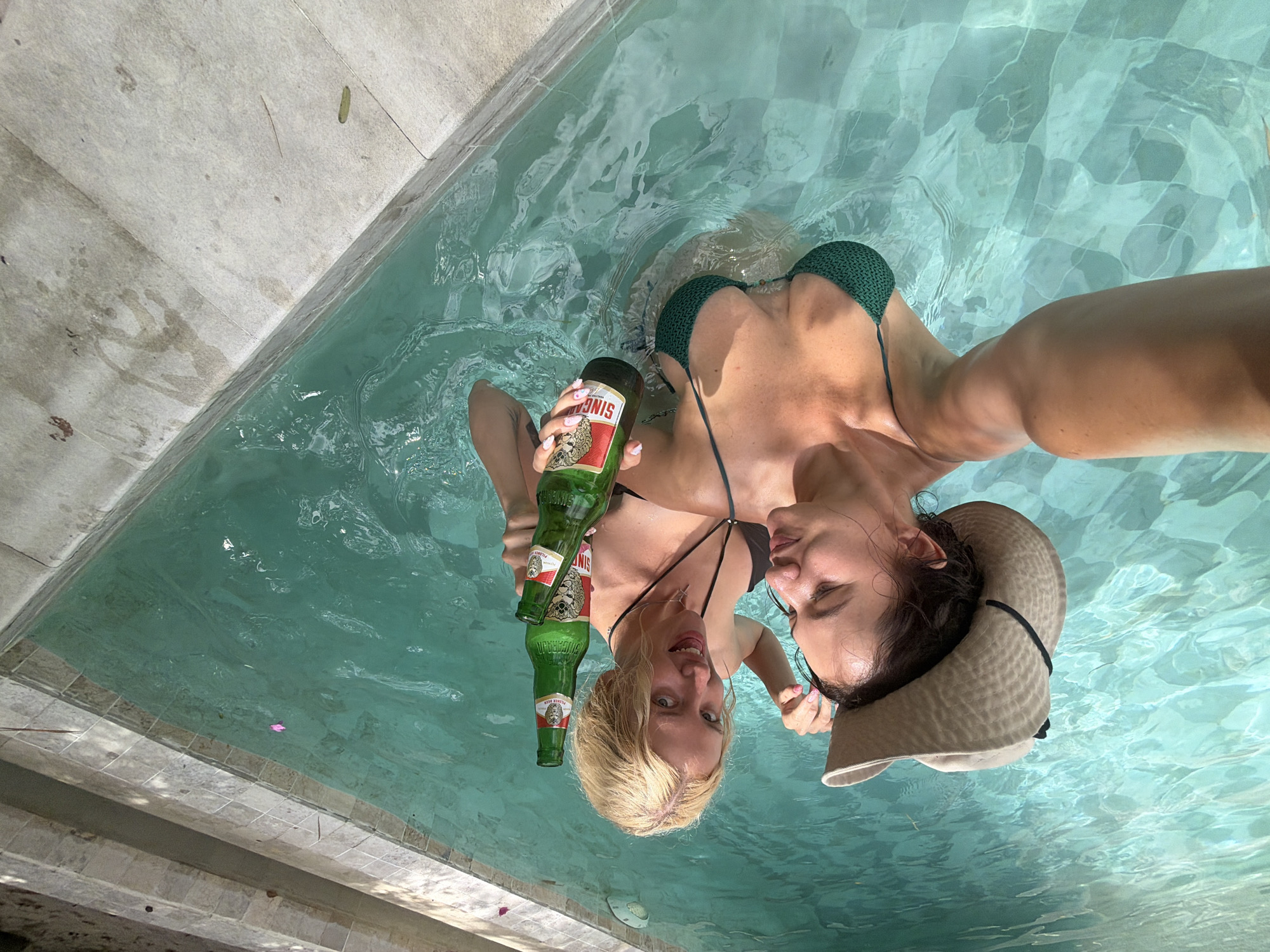 Two women are in a swimming pool in North Kuta, Indonesia. One woman is wearing a wide-brimmed hat and a green bikini top. The other woman is wearing a black bikini top. Both women are holding green glass bottles of beer. The pool is surrounded by light-colored tile and the water is clear and turquoise.
