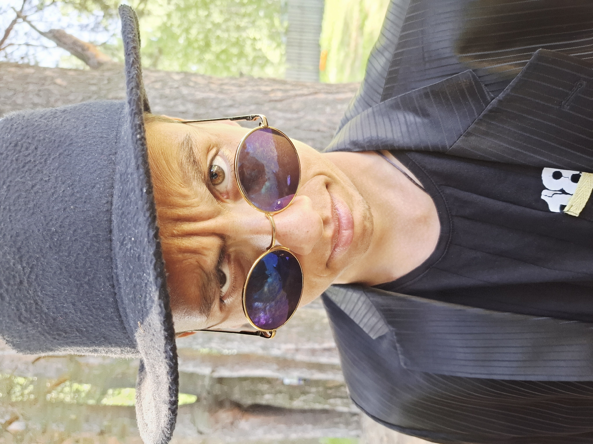 A man is shown from a slightly angled, close-up perspective. He is wearing a dark gray or black felt hat, round sunglasses with iridescent lenses and gold frames, a black t-shirt with a white graphic, and a dark pinstripe jacket. The background is blurred, suggesting an outdoor location with greenery, possibly a park. A log or branch is visible behind the man. It cannot be determined from the image if the location is in Chaikovsky, Russia.