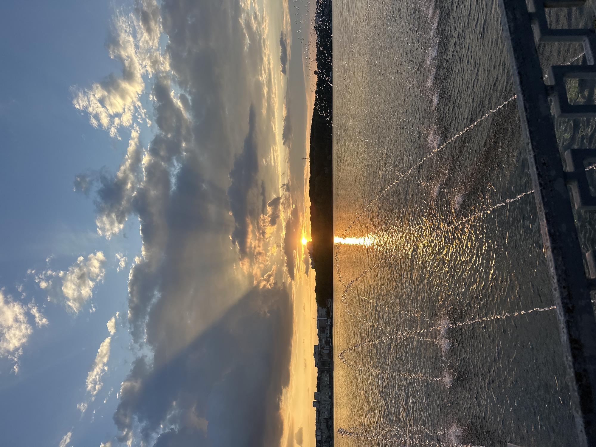 A wide shot captures a sunset over a body of water in Ternopil, Ukraine. The sun, partially obscured by clouds, casts a golden glow across the sky and reflects on the water's surface. Light rays emanate from behind a dark cloud formation on the left, creating a dramatic effect. In the background, a cityscape with buildings and a line of trees along the horizon can be seen. In the foreground, water jets from a fountain create arcs of spray across the water. A metal railing is visible in the bottom foreground, suggesting the vantage point is from a bridge or promenade. The water is rippled by the fountain's activity and the natural movement of the water.