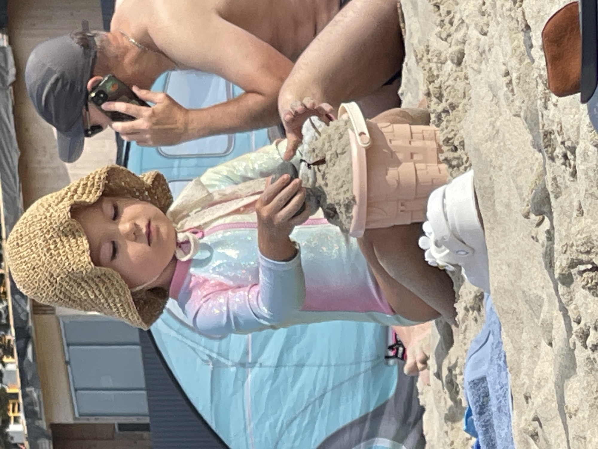 The image depicts a beach scene with two individuals and assorted objects. In the foreground, a young child, wearing a straw hat and a colorful swimsuit, is seated in the sand, actively playing with a pink sand bucket and sand. A white sandal is visible to the child's right. Behind the child, an adult male, wearing a grey baseball cap and sunglasses, is seen talking on a mobile phone. He is shirtless, exposing his upper body. In the background, a blue tent or beach shelter is partially visible. The environment is sandy, characteristic of a beach. The individuals appear to be at a beach location, possibly in Zelenogradsk, Russia, given the context of the prompt.